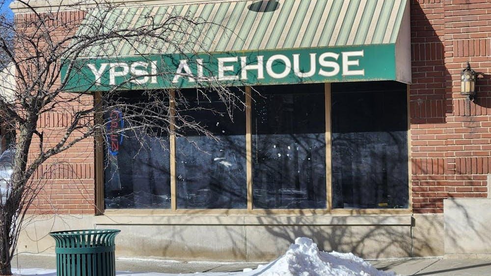 The exterior of a red brick building with a green awning over a large window. The awning says "Ypsi Alehouse" in capital white letters. In front of the building are a pile of white snow and a tree that has lost all its leaves.