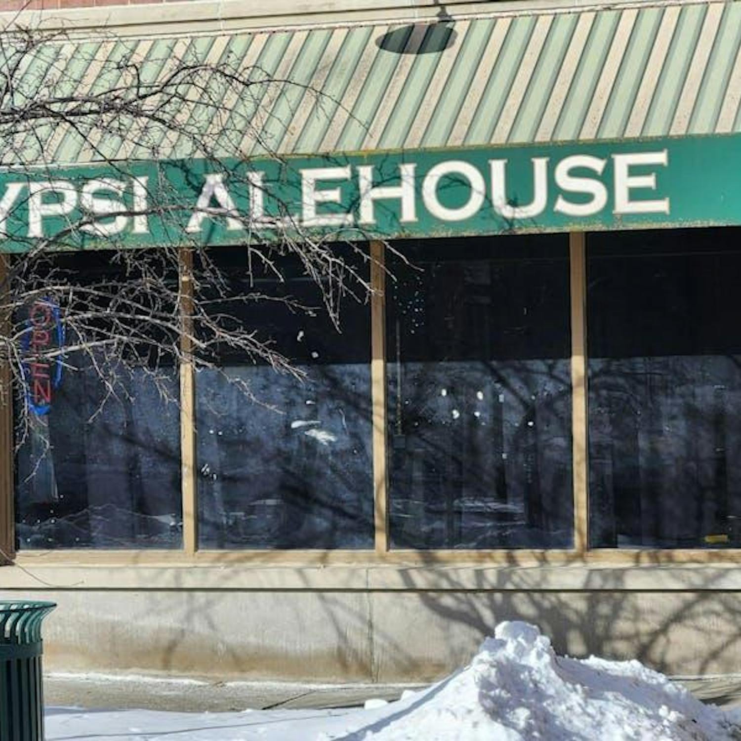 The exterior of a red brick building with a green awning over a large window. The awning says "Ypsi Alehouse" in capital white letters. In front of the building are a pile of white snow and a tree that has lost all its leaves.