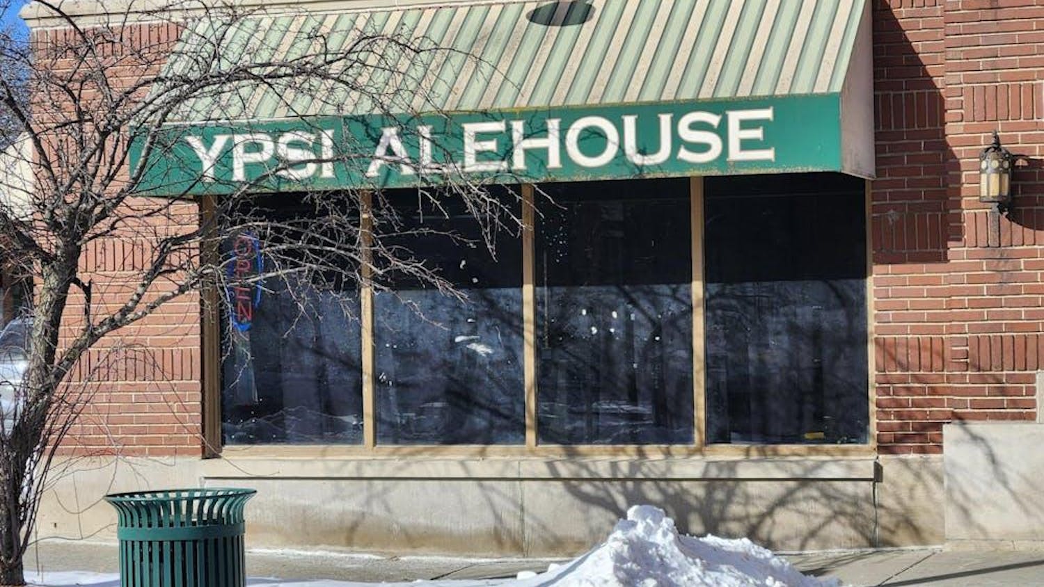 The exterior of a red brick building with a green awning over a large window. The awning says "Ypsi Alehouse" in capital white letters. In front of the building are a pile of white snow and a tree that has lost all its leaves.