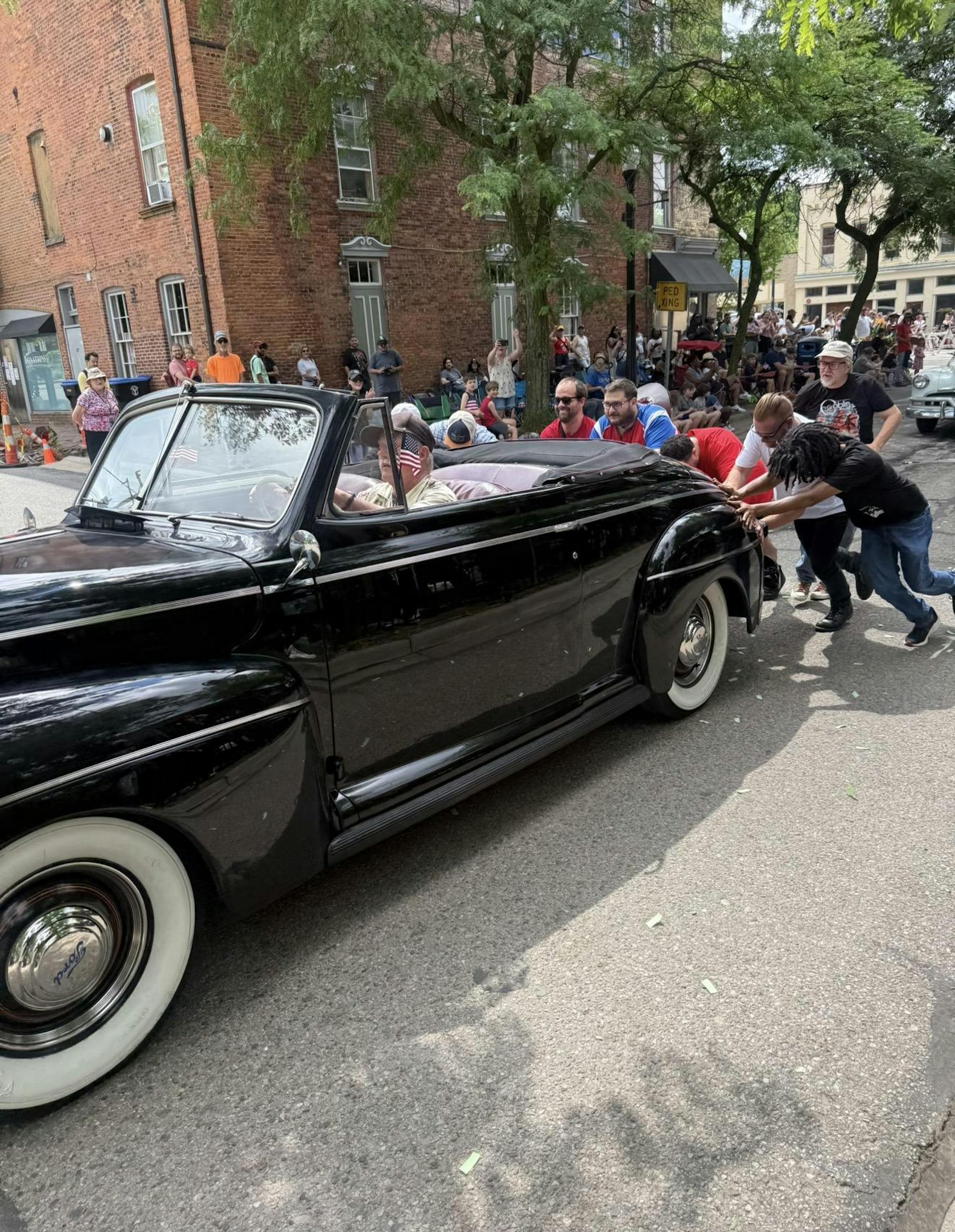 People push a vintage car through the streets while a man drives.