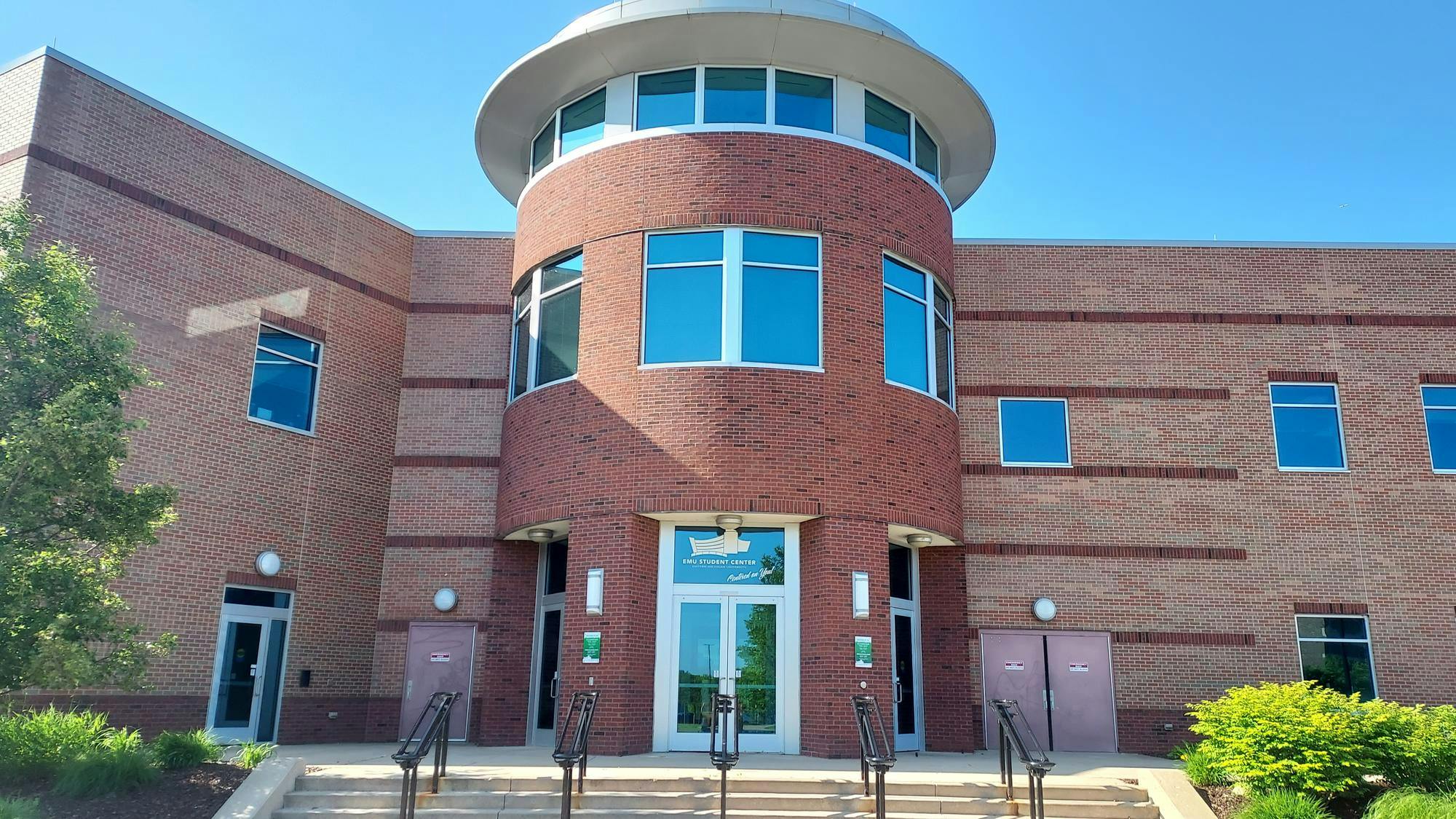 The entrance to the EMU Student Center is via several concrete steps. The Student Center exterior is made of red brick, and its glass doors are framed.