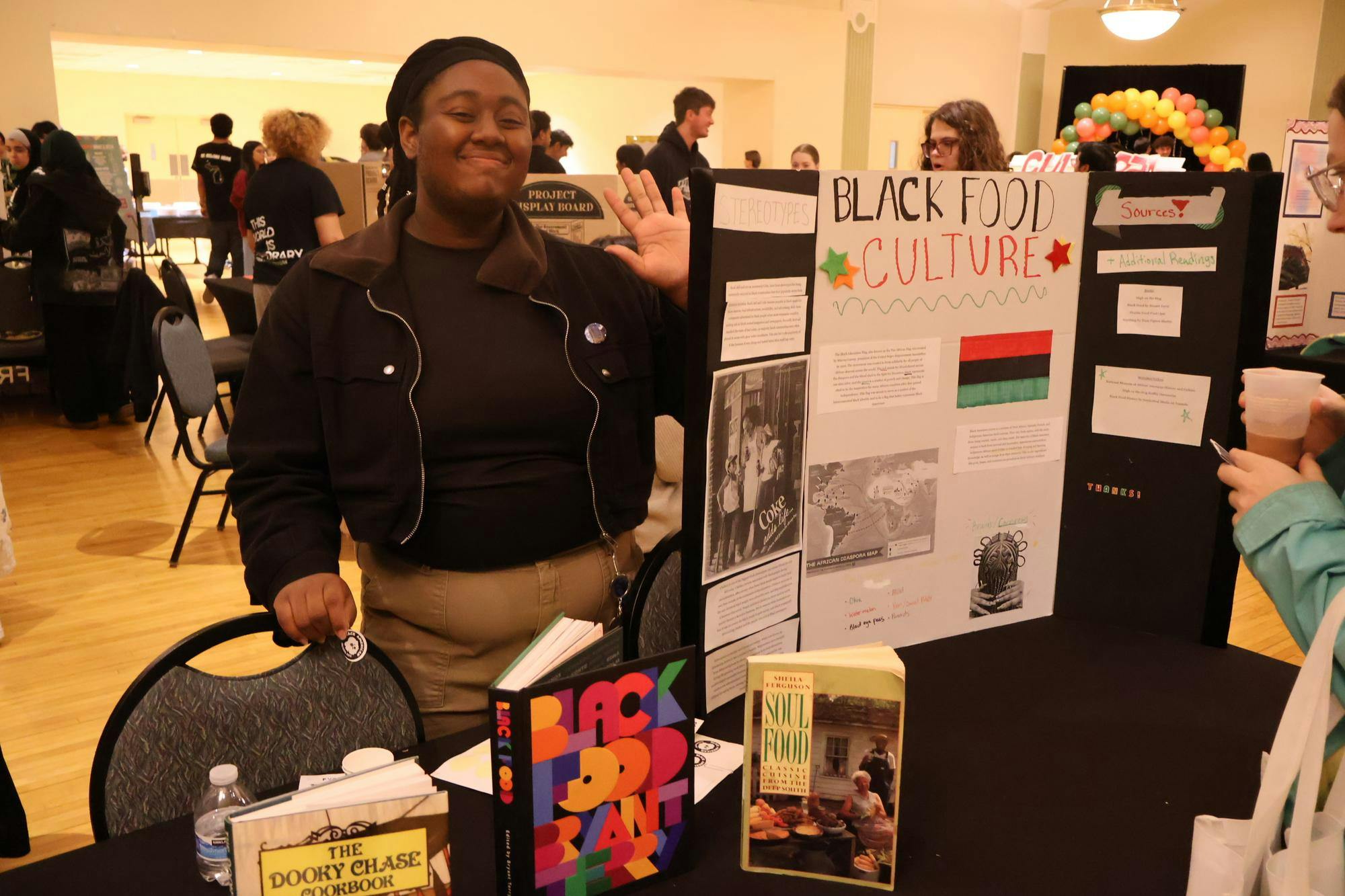 A student waves to the camera while standing next to their poster board.
