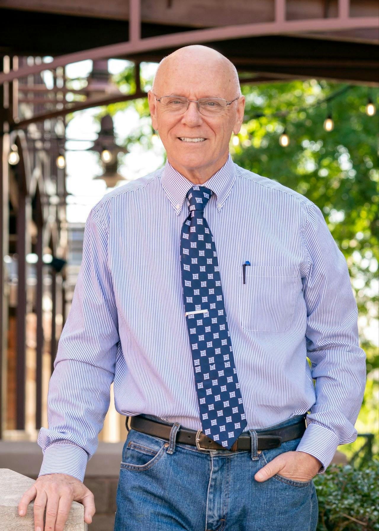 A man wearing a blue and white striped shirt with a dark blue tie stands outside to pose for a photo.