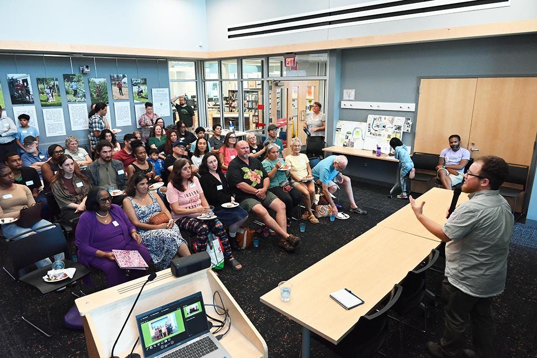 A crowd sits indoors on chairs and watches a speaker.