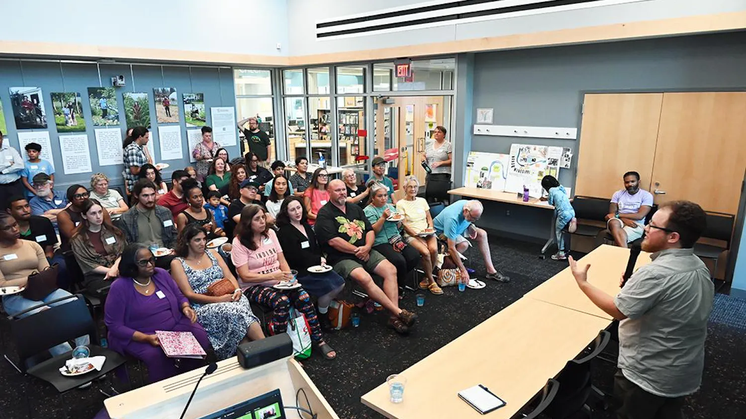 A crowd sits indoors on chairs and watches a speaker.