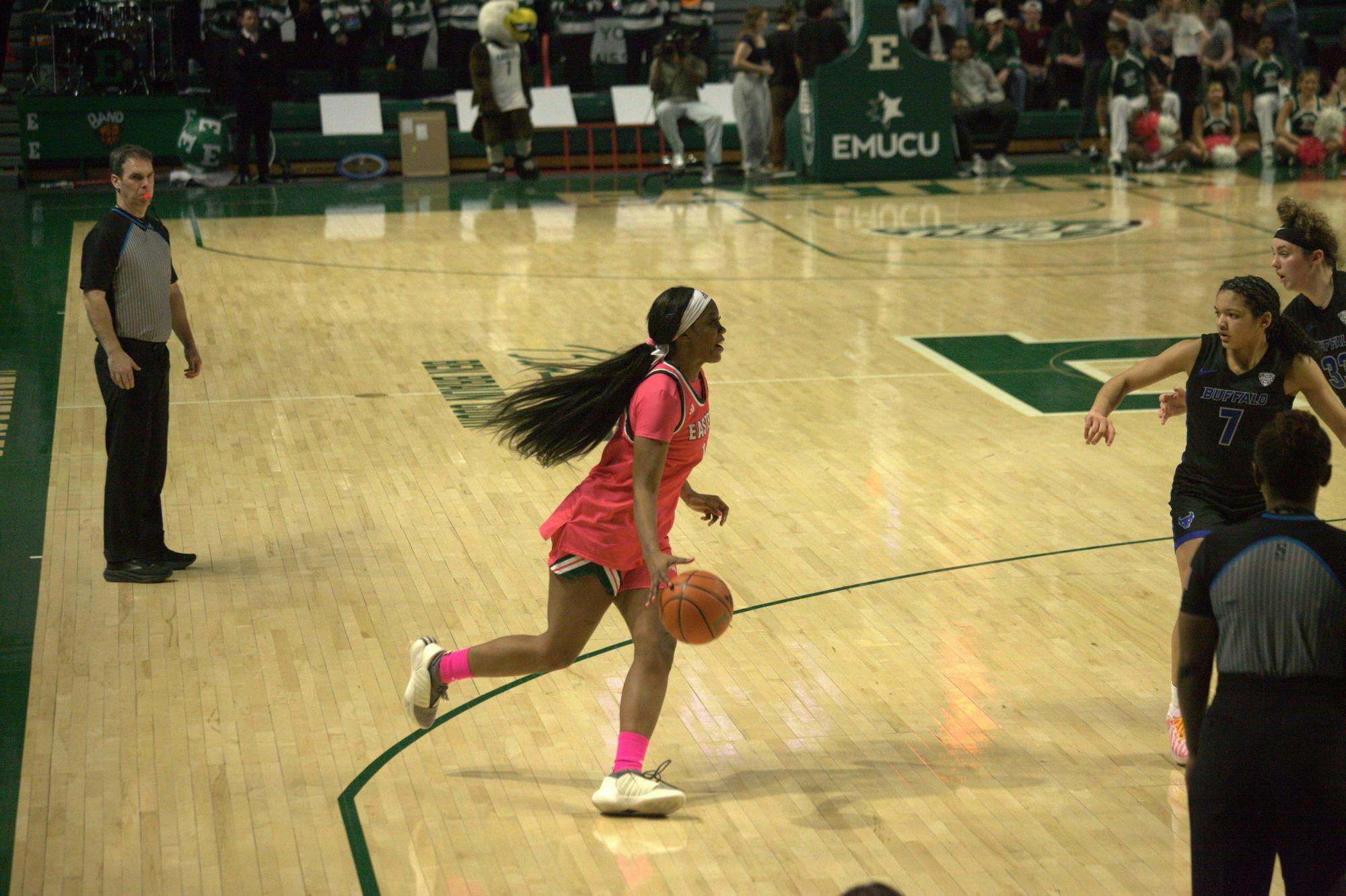 A women's basketball player dribbles the ball across the court towards the opposing team's hoop while referees and members of the opposing team watch.