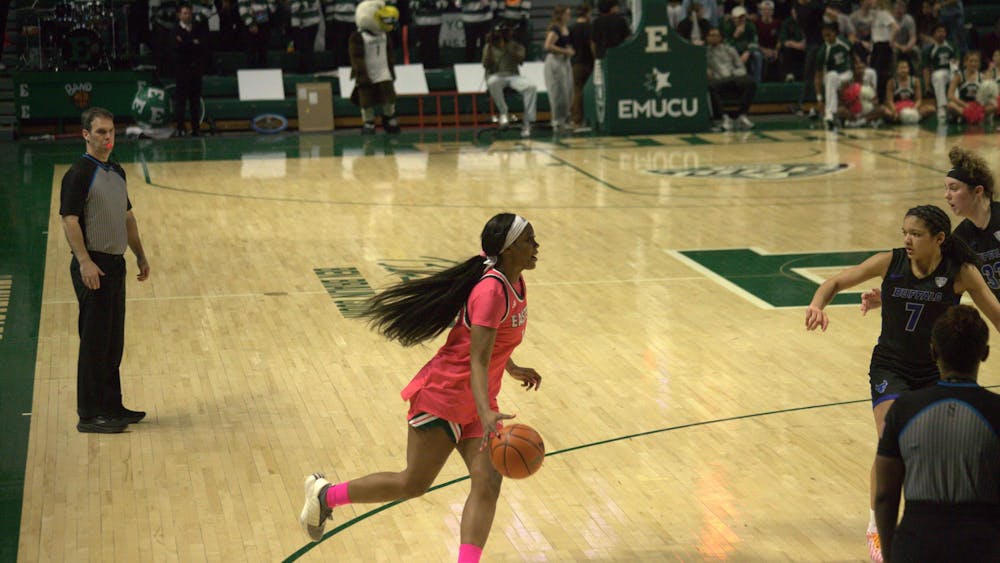 A women's basketball player dribbles the ball across the court towards the opposing team's hoop while referees and members of the opposing team watch.