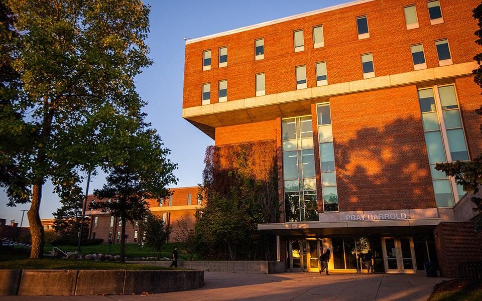 Students walk outside of a large red brick building while the sun sets.