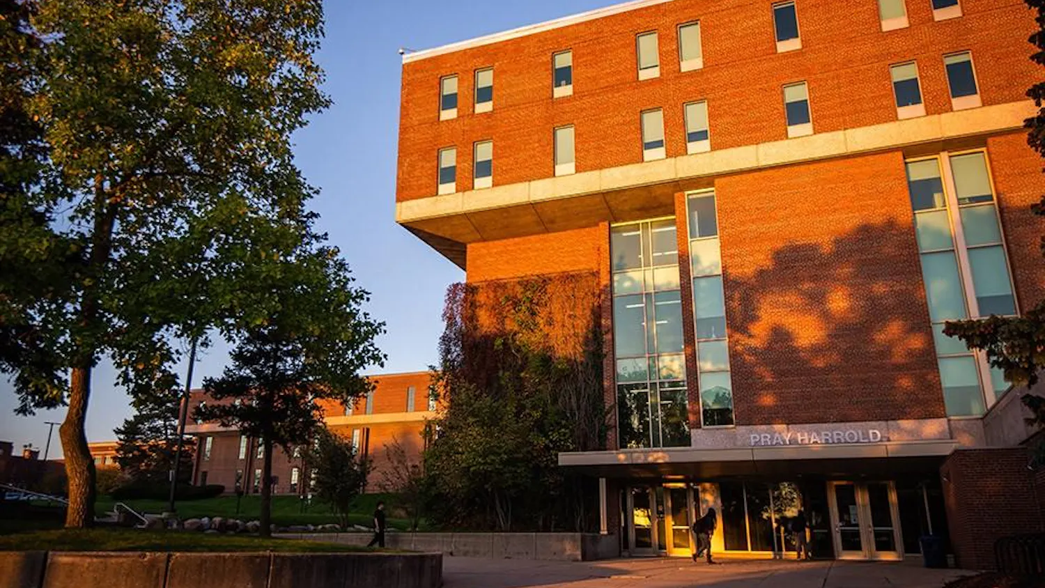 Students walk outside of a large red brick building while the sun sets.