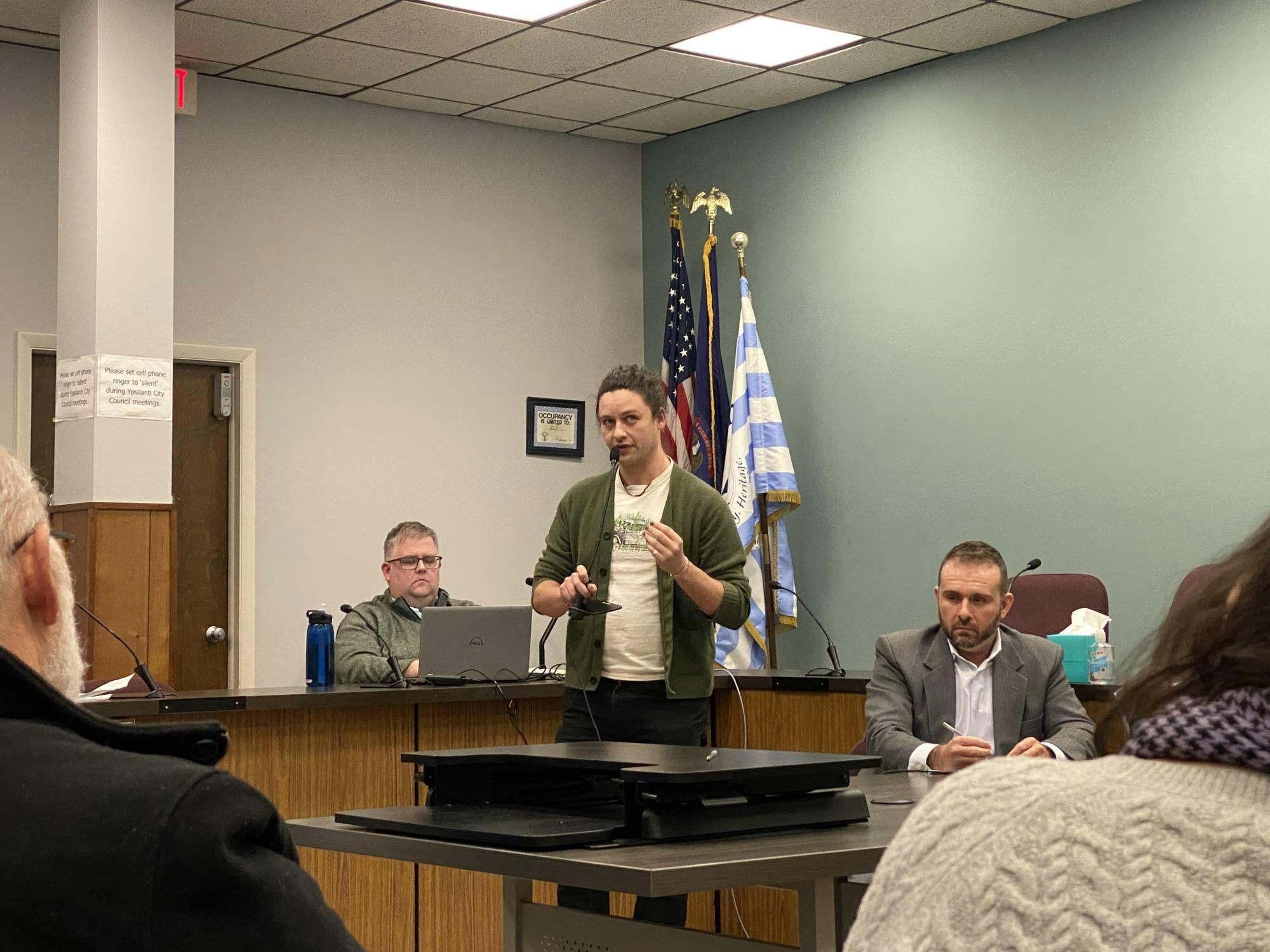 Brian Geiringer, center, speaks into a microphone. Two people are seated behind him at a desk and table.