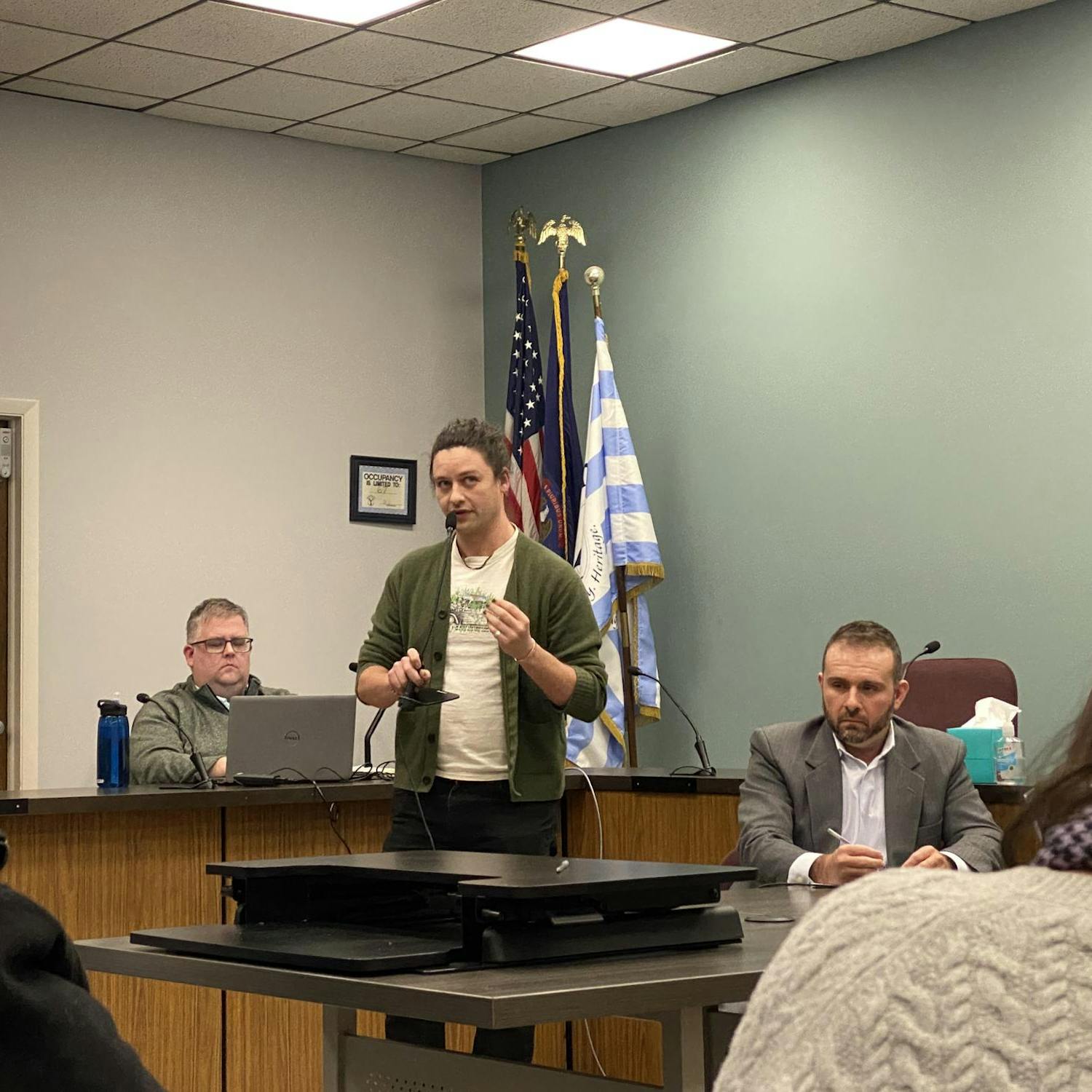 Brian Geiringer, center, speaks into a microphone. Two people are seated behind him at a desk and table.