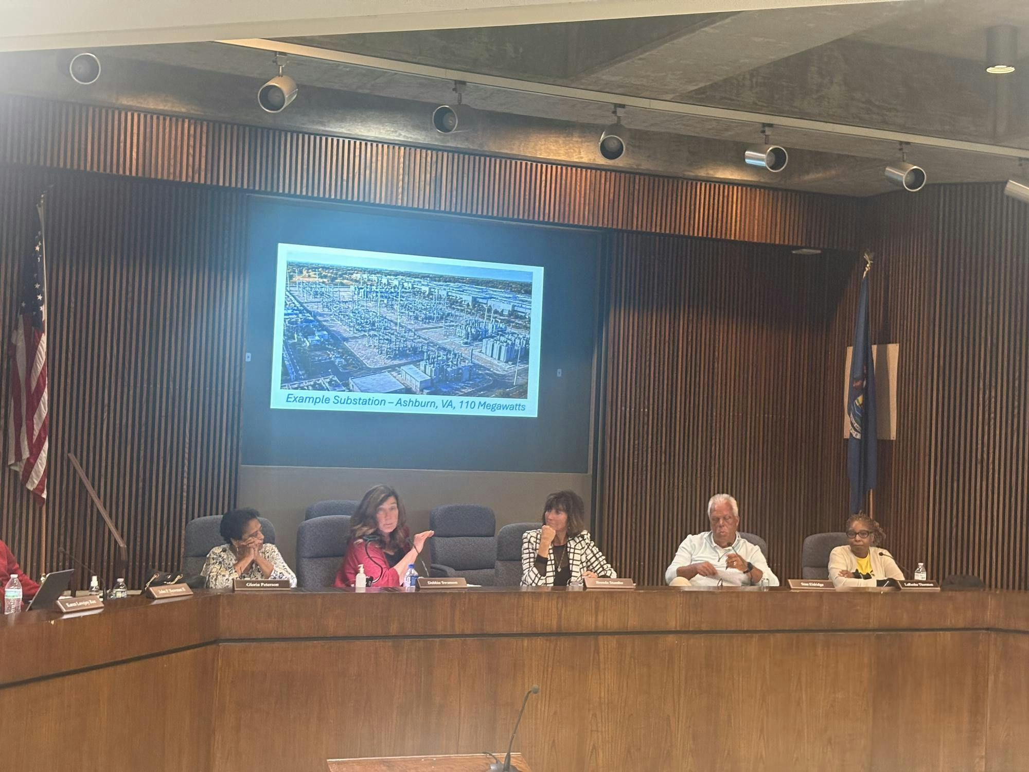 Five people sit at a wooden desk in the Ypsilanti Township civic center meeting room. Behind them, a projector shows an example of a 110-megawatt substation.