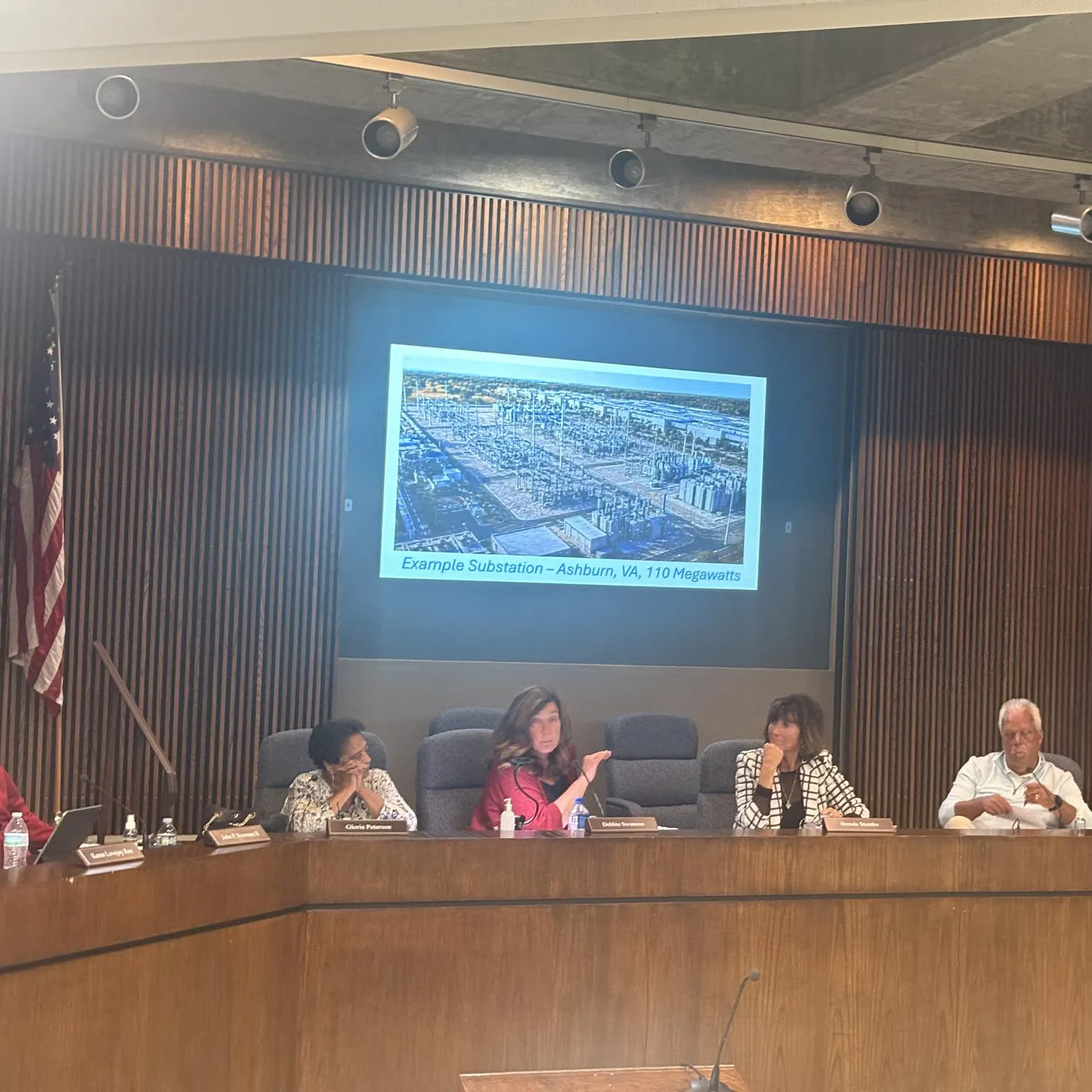 Five people sit at a wooden desk in the Ypsilanti Township civic center meeting room. Behind them, a projector shows an example of a 110-megawatt substation.