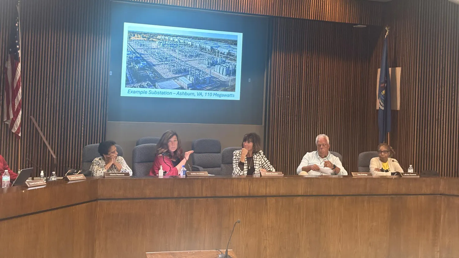 Five people sit at a wooden desk in the Ypsilanti Township civic center meeting room. Behind them, a projector shows an example of a 110-megawatt substation.