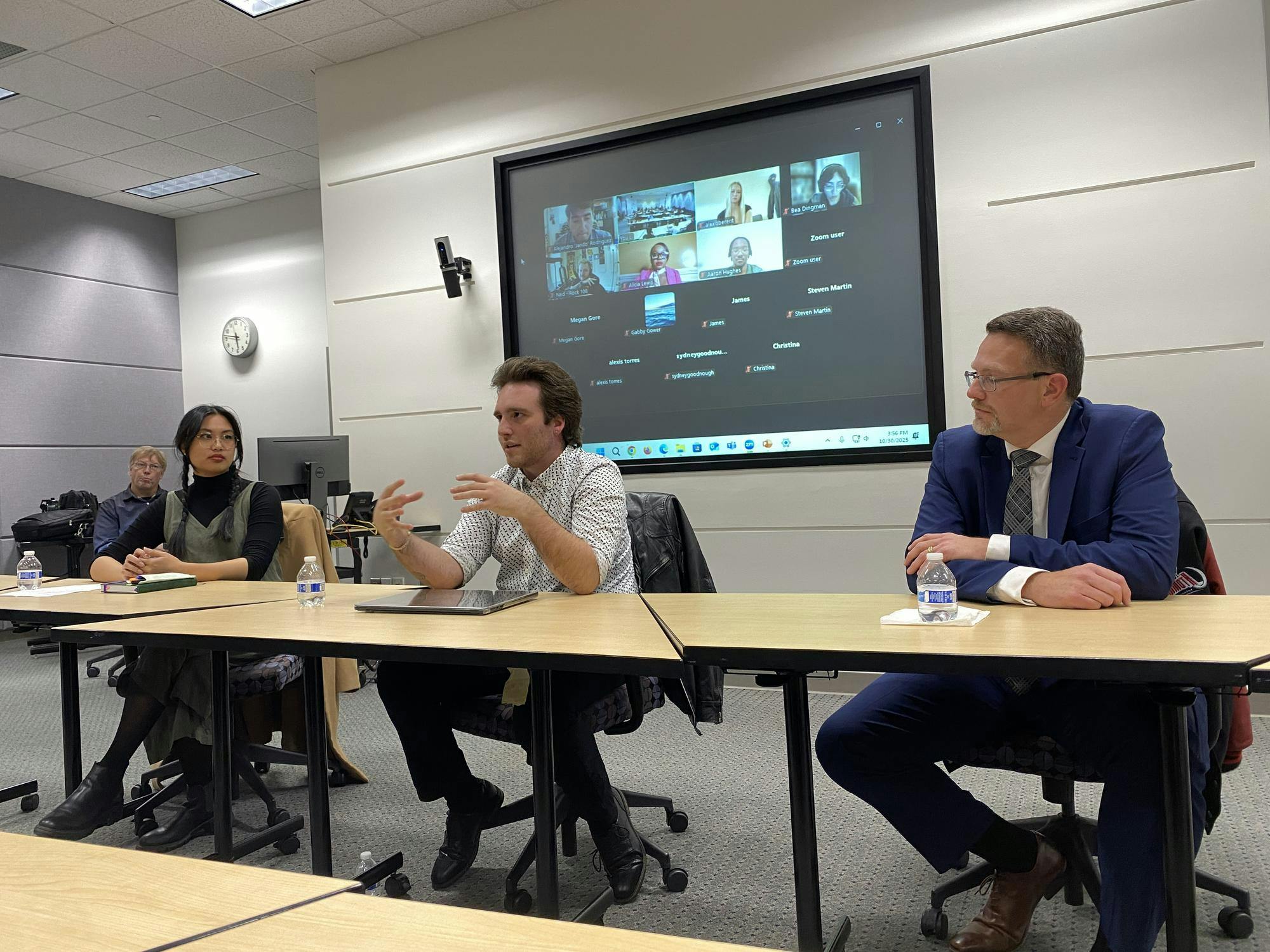 Three alumni panelists sit at a long table. The panelists on either end turn to listen to the middle panelist speak. A large screen in the background shows participants listening through Zoom.