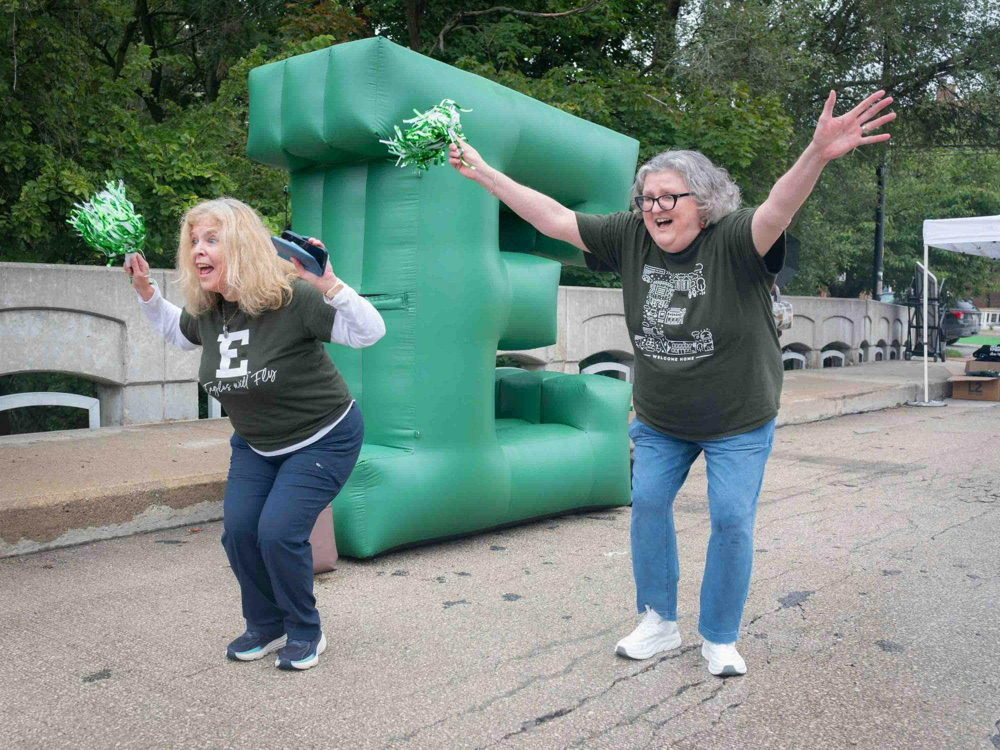Jackie LaRose and Wendy Burke smile and wave green and white pom-poms while standing in front of a giant inflatable green block letter E.