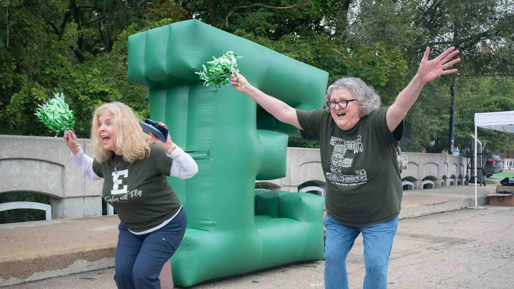 Jackie LaRose and Wendy Burke smile and wave green and white pom-poms while standing in front of a giant inflatable green block letter E.