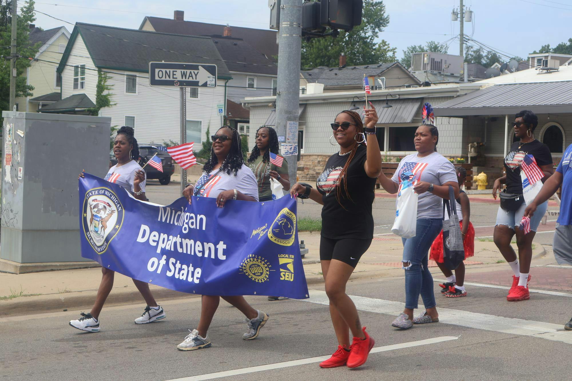 Multiple African Americans wearing shirts with American flags on them holding a sign that says "Michigan Department of State". Many are smiling and waving miniature American flags. 