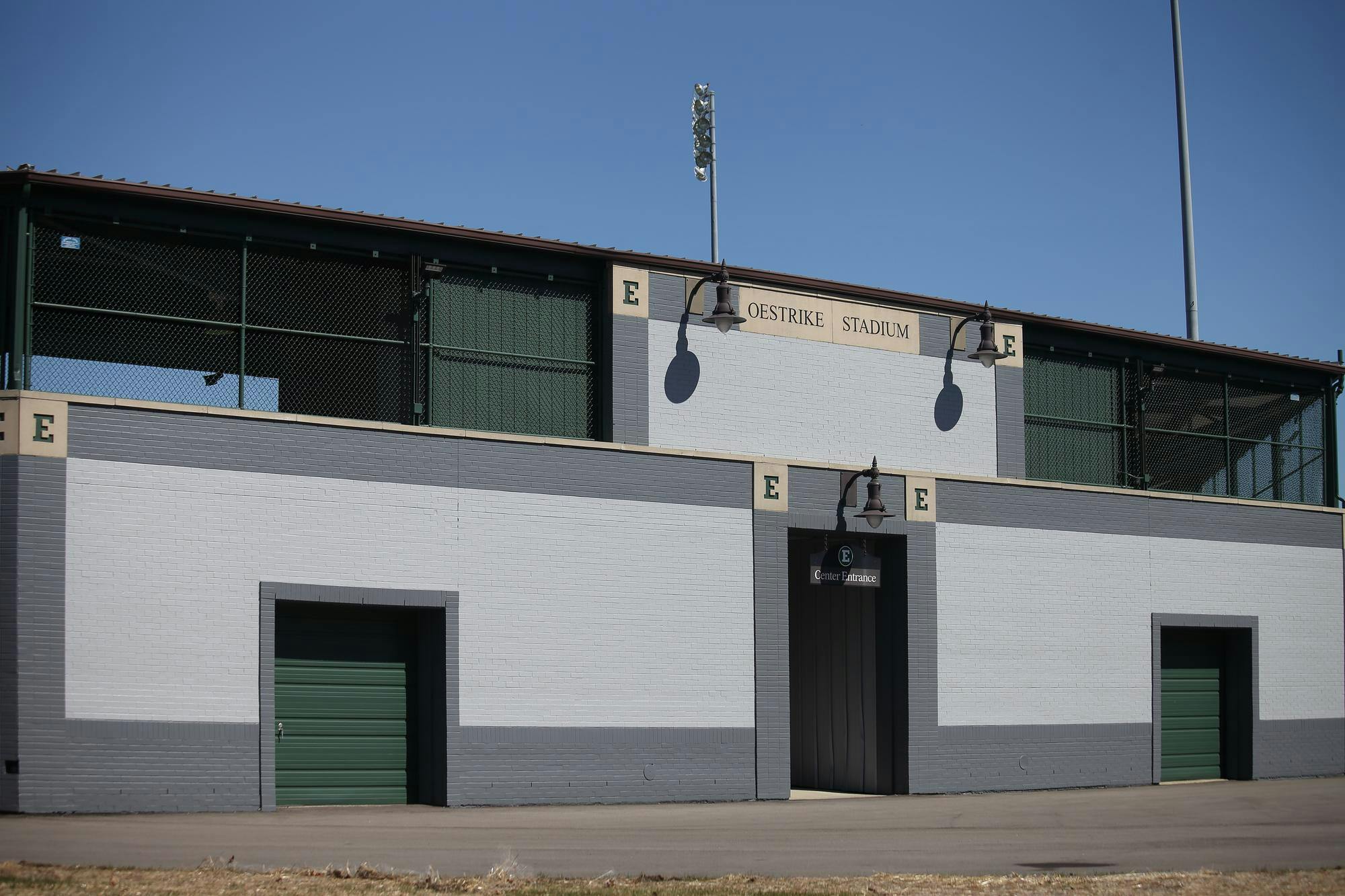 The wall of a baseball stadium from outside. Text in the top center reads "Oestrike Stadium." One entrance is visible with a hanging sign that reads "Center Entrance."