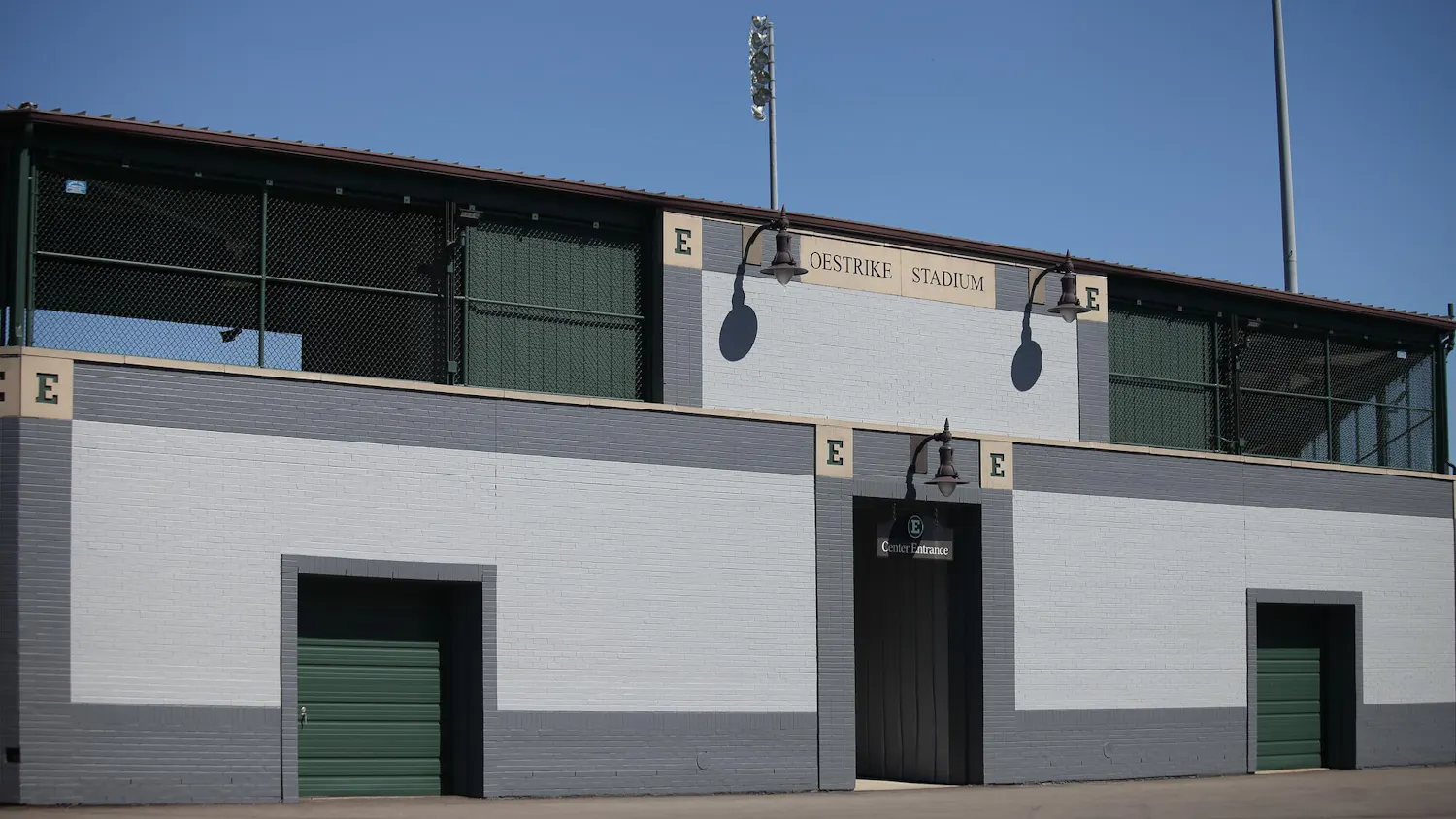 The wall of a baseball stadium from outside. Text in the top center reads "Oestrike Stadium." One entrance is visible with a hanging sign that reads "Center Entrance."