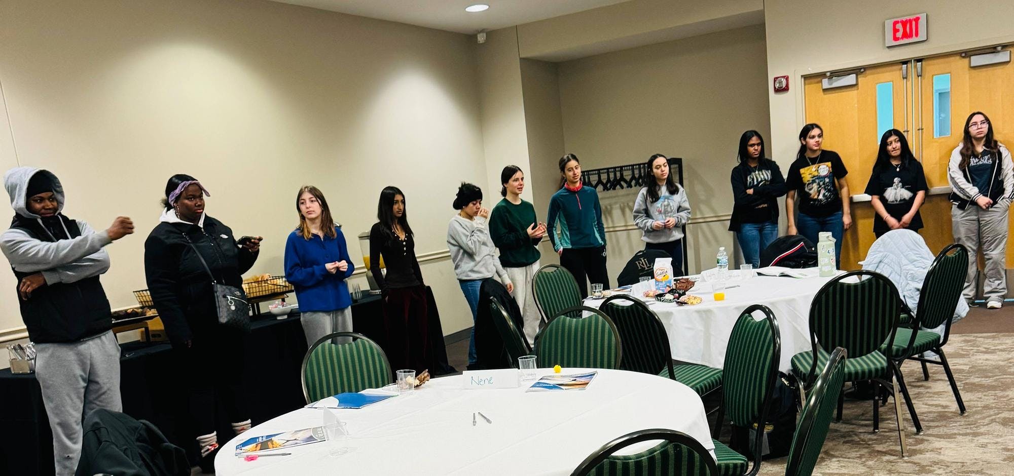 Several middle school students stand in a room with beige walls, and four of the teens are standing in front of a black table with snacks. In front of the teens are round tables with white tablecloths and dark green striped chairs. 