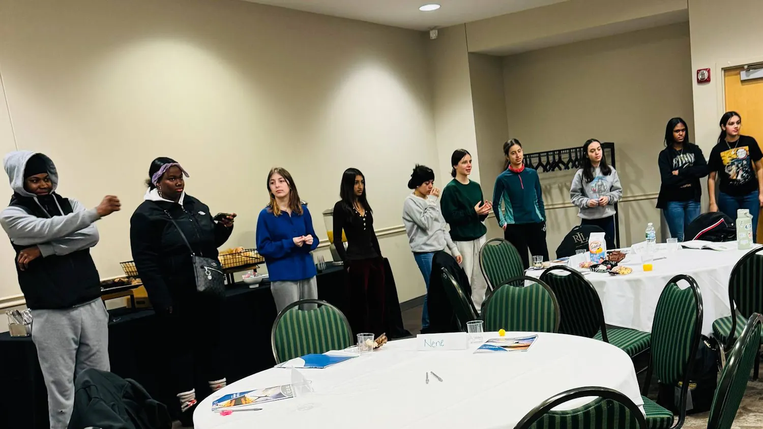 Several middle school students stand in a room with beige walls, and four of the teens are standing in front of a black table with snacks. In front of the teens are round tables with white tablecloths and dark green striped chairs.