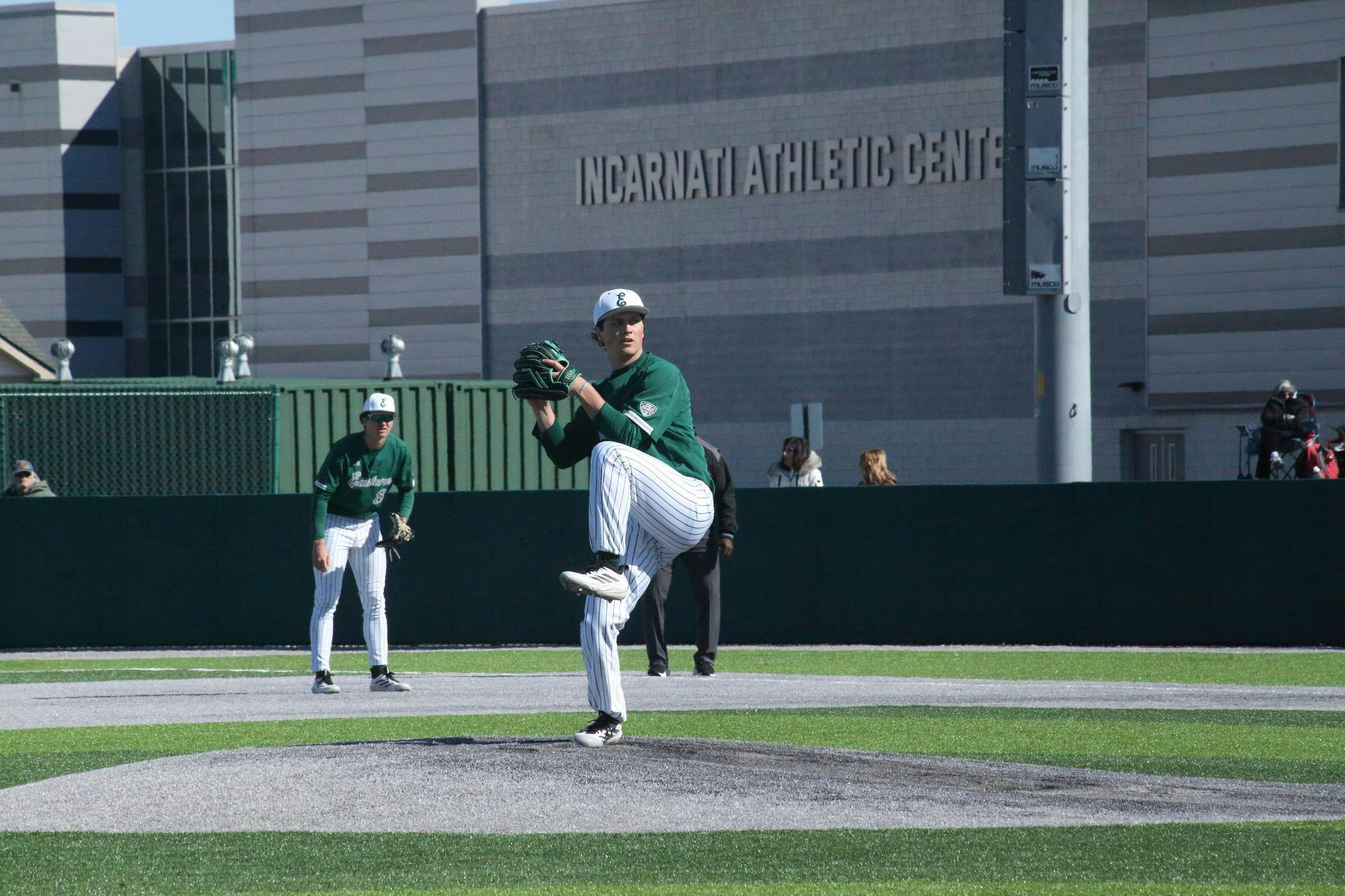 A pitcher winds up in preparation to pitch the ball. A teammate and an umpire are visible in the background on the field.