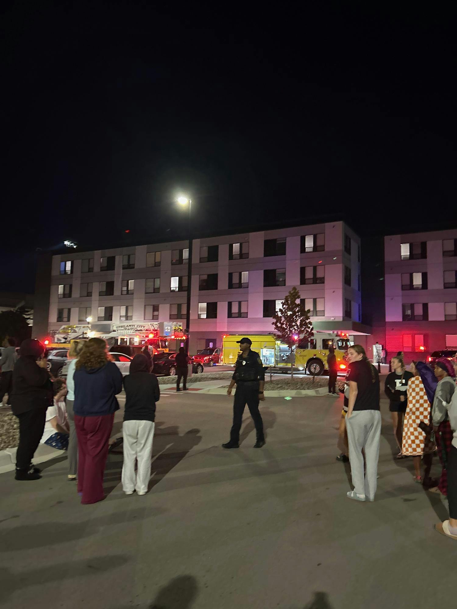 Students gather outside and face the gray Westview Apartments building. Two fire trucks are parked in front of the apartments. Police and firefighters arrive on scene to manage the fire.
