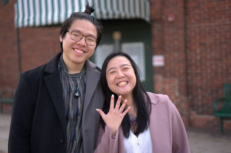 Christopher Kok and Julian Ho stand beside one another smiling while Julian Ho holds up her hand to show off her new engagement ring. 