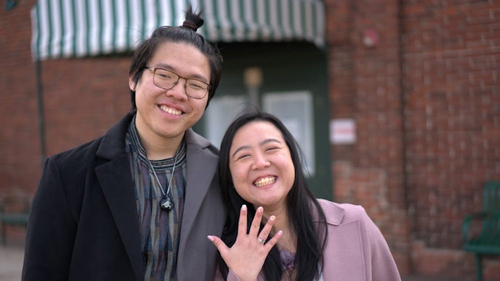 Christopher Kok and Julian Ho stand beside one another smiling while Julian Ho holds up her hand to show off her new engagement ring.