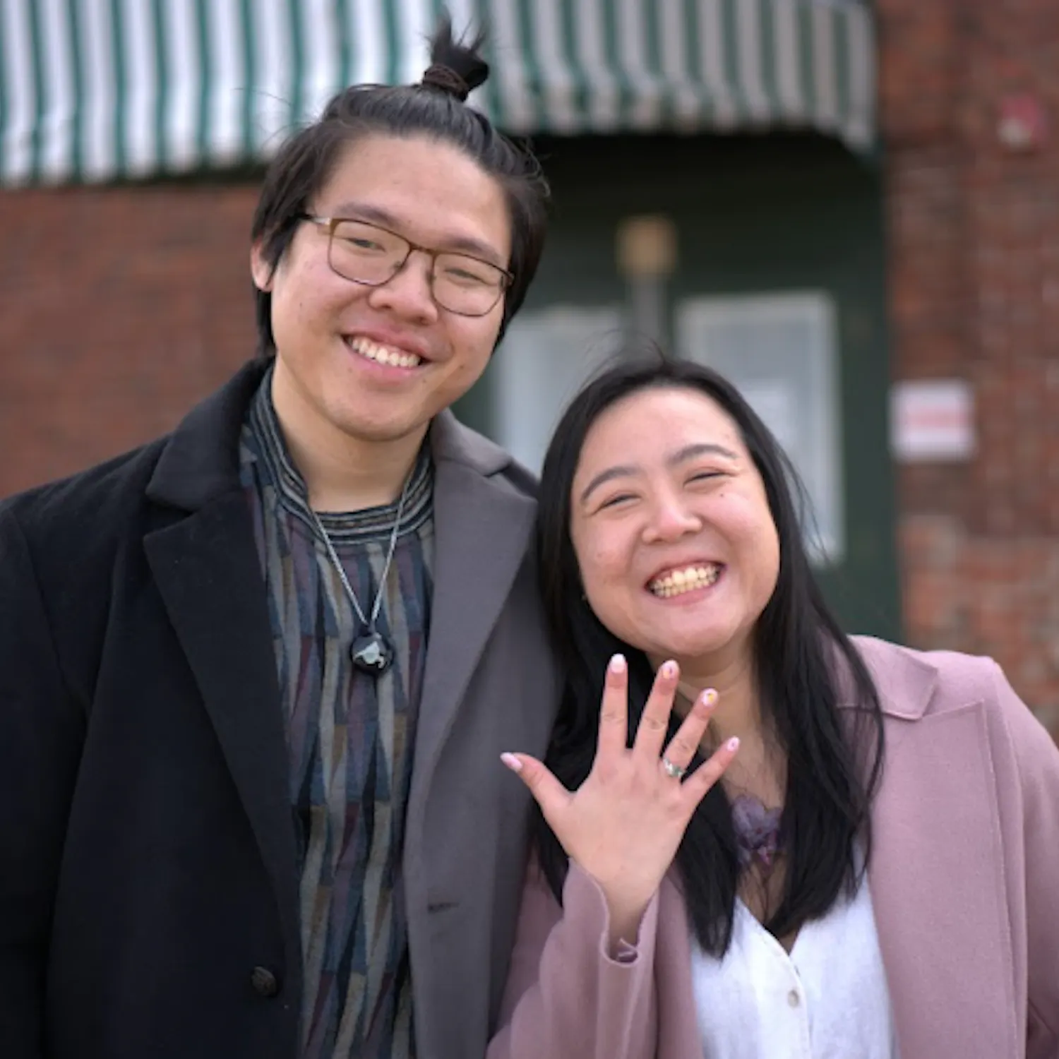 Christopher Kok and Julian Ho stand beside one another smiling while Julian Ho holds up her hand to show off her new engagement ring.