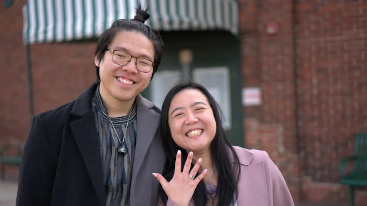 Christopher Kok and Julian Ho stand beside one another smiling while Julian Ho holds up her hand to show off her new engagement ring.