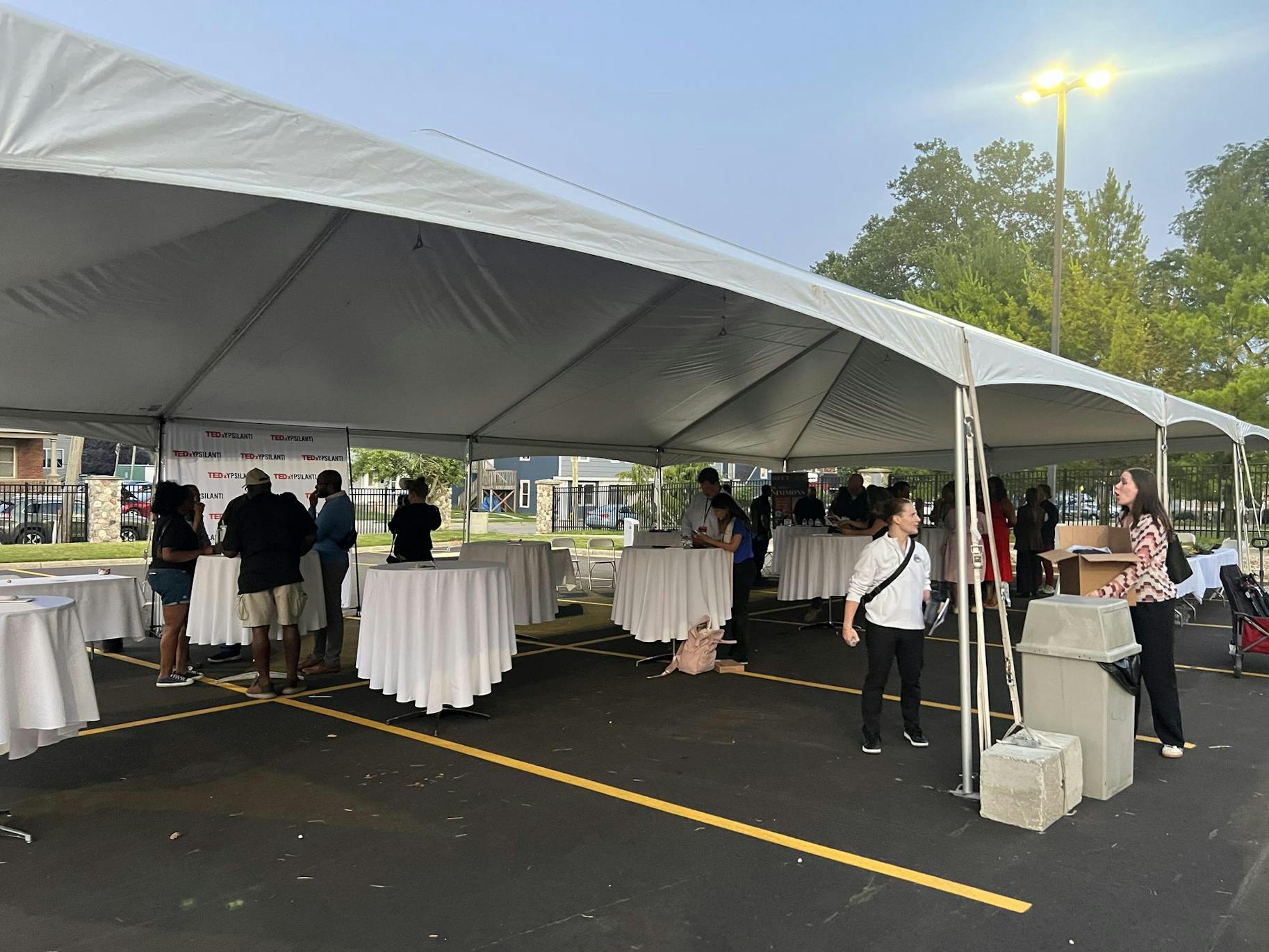 Dwindling groups of people stand at tables under a canopy in a parking lot. There is a TEDxYpsilanti photo wall. 
