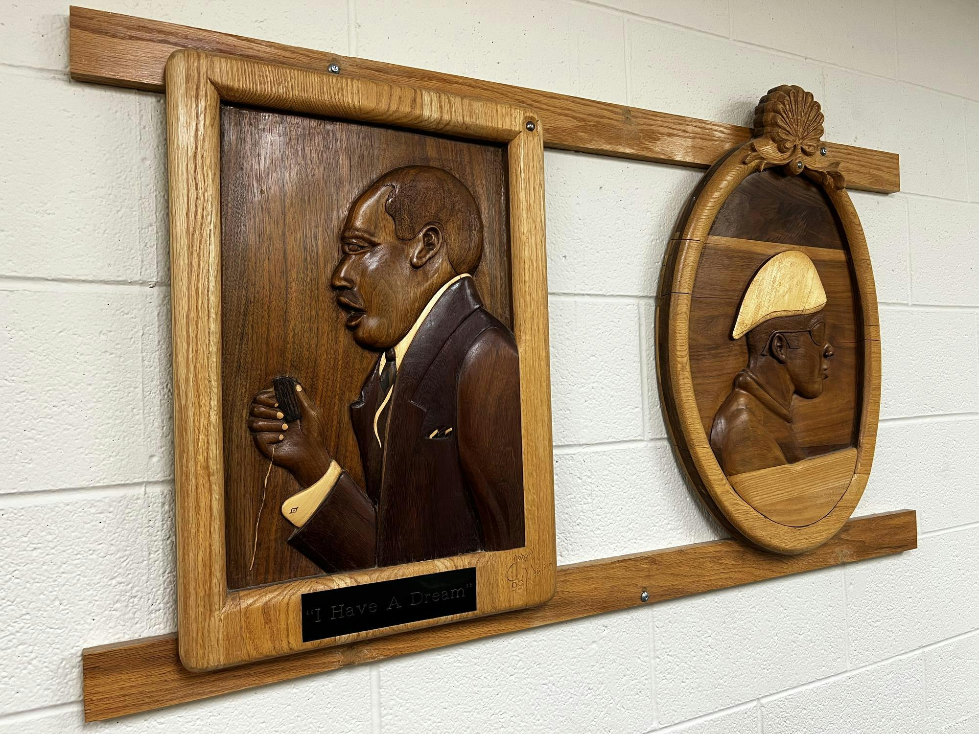 Two engraved wooden portraits on a beige brick wall. Under Rev. Dr. Martin Luther King Jr. is the quote, "I have a dream." The man depicted in the second artwork is wearing sunglasses and a backwards cap.