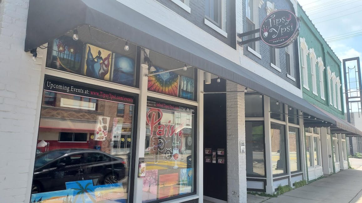 The side of a windowed building with a circular sign reading "Tipsy Ypsi" on top, with multiple colorful paintings displayed in the window.