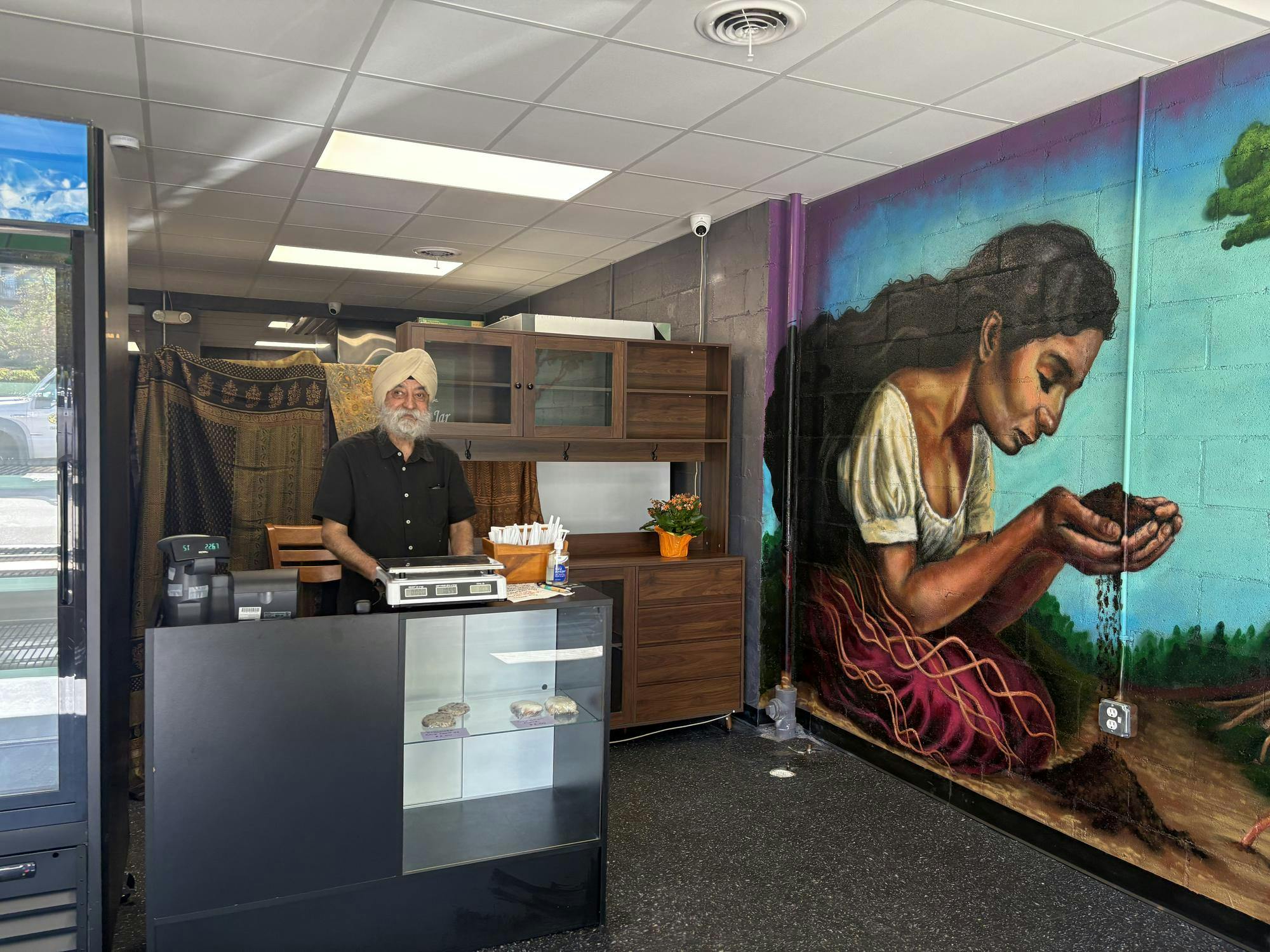Pushpinder Sethi stands behind the register counter in front of brown cabinets and a tapestry. A mural is painted on the wall next to him of a woman kneeling on the ground and holding dirt in her hands, suggesting a spiritual connection to the Earth.