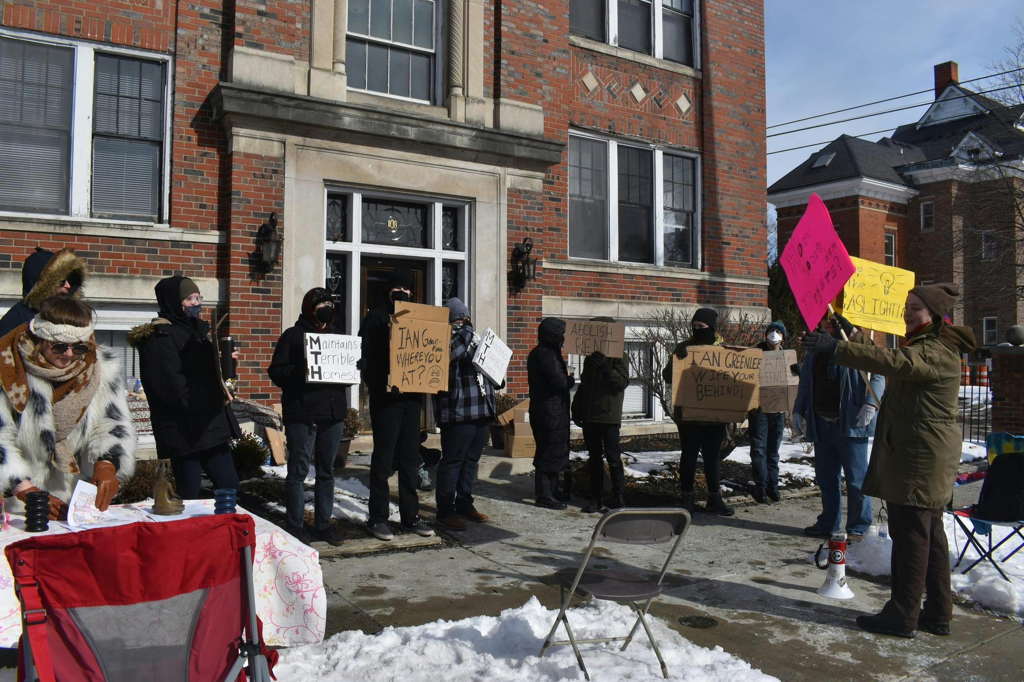 10 people gather on the sidewalk in front of a brick apartment building. They hold signs in protest of the apartment's conditions.