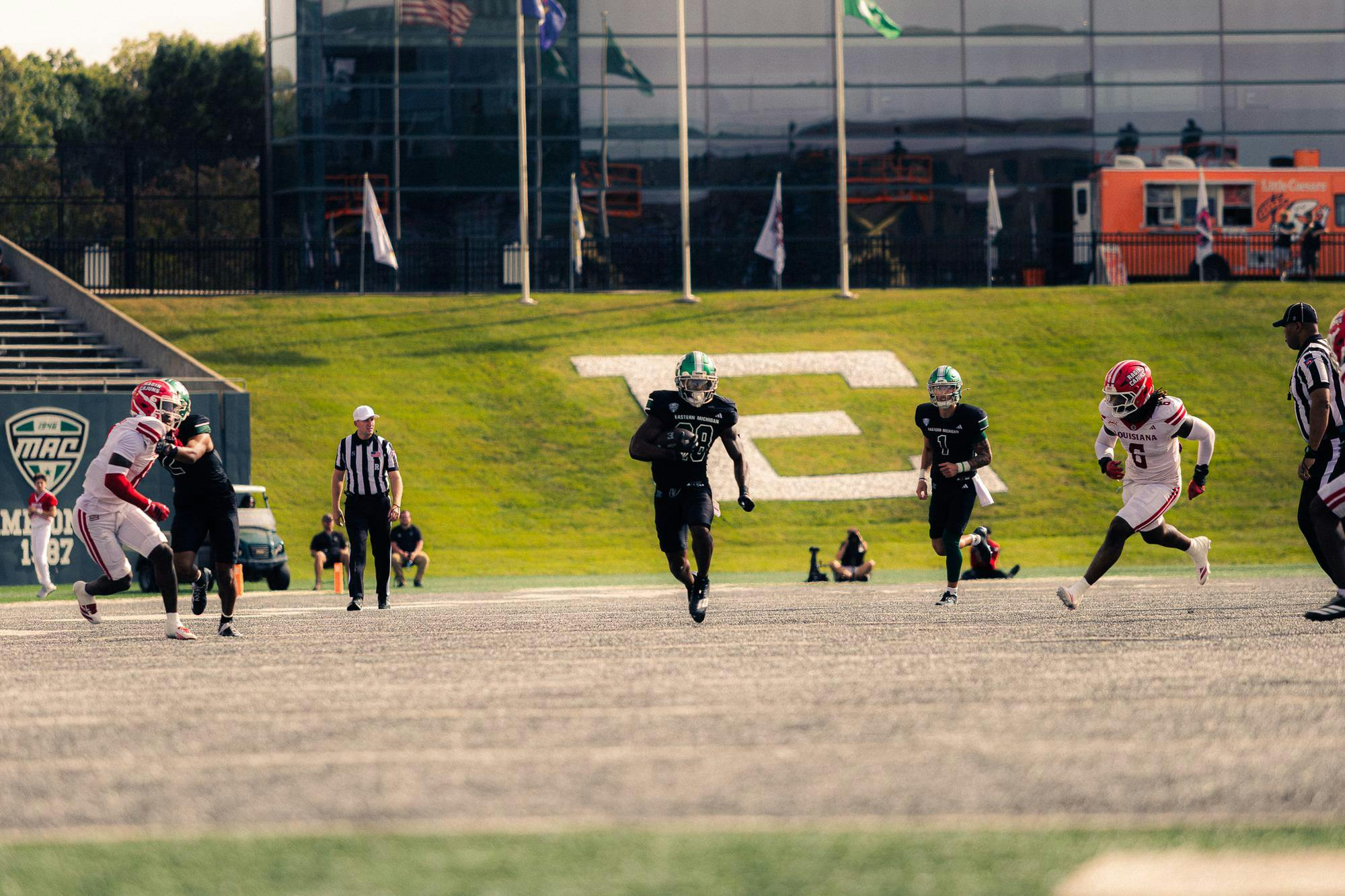 An Eastern Michigan football player wearing green and black with a green helmet carries the ball into open space on the gray grass of the football field in front of him.