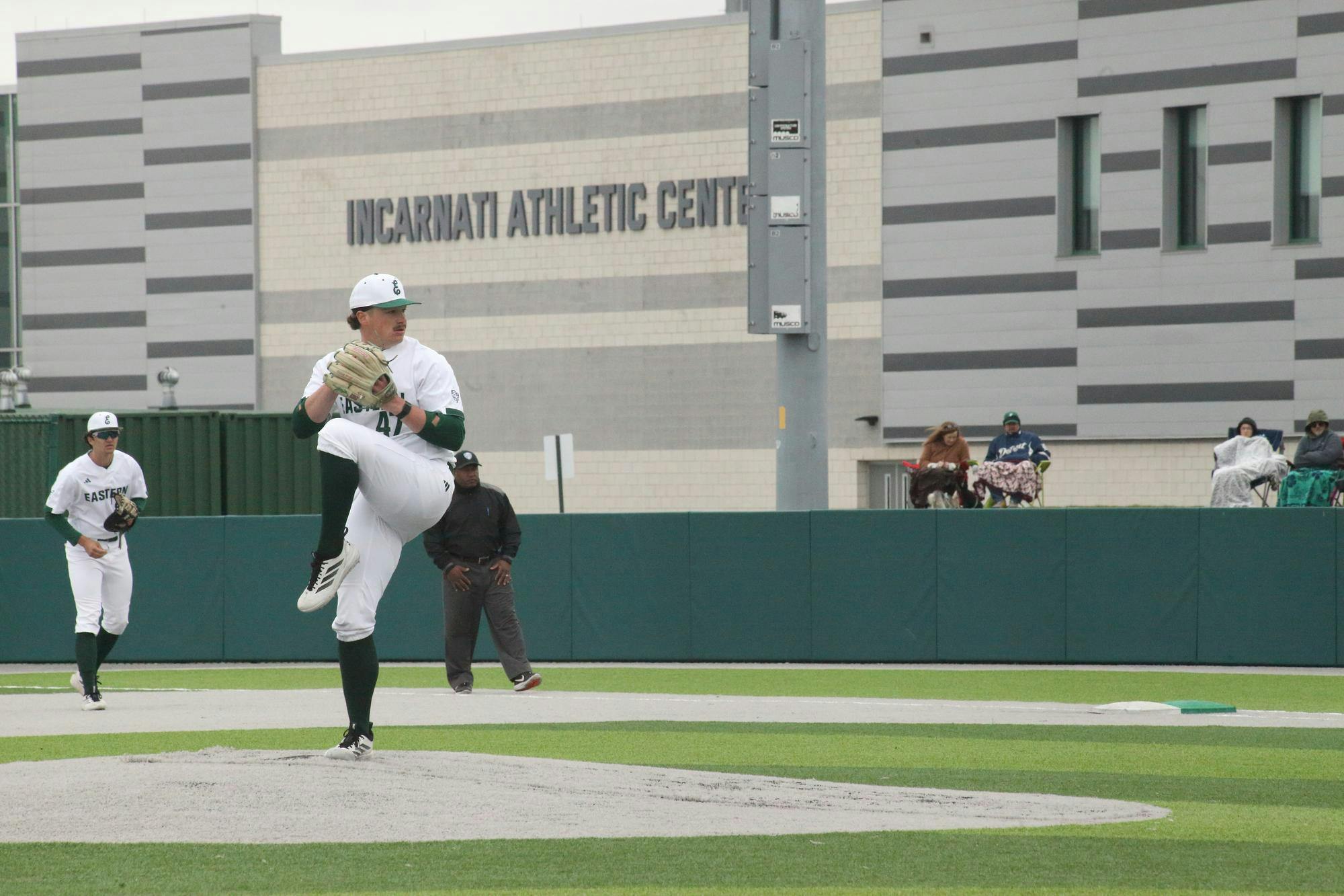 Baseball player winds up pitch while teammate, umpire, and spectators watch at varying distances.