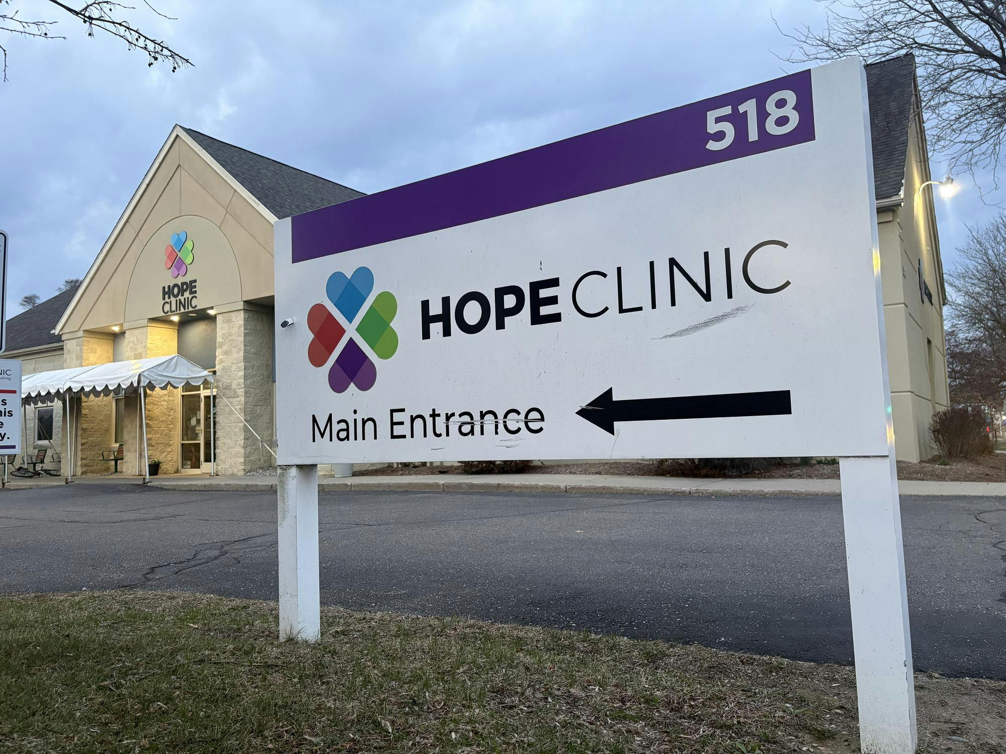 White and purple outdoor sign reading, "Hope Clinic, Main Entrance." The beige concrete and brick building of Hope Clinic is in the background.