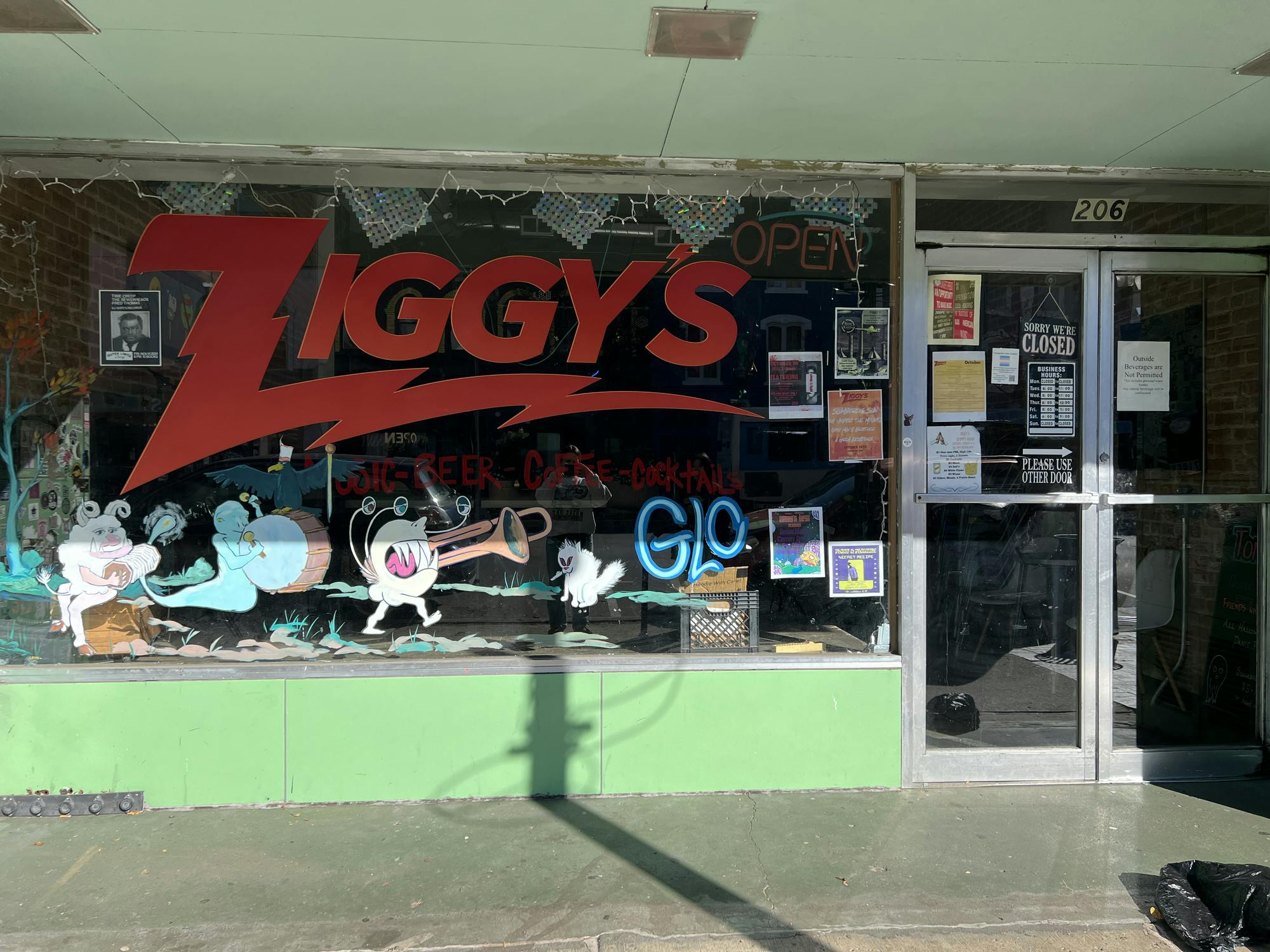 A storefront with the word "Ziggy's" written in big red letters on the building's glass wall. Underneath the writing are several colorful drawings of mythical creatures, some of them playing musical instruments. Off to the right are two glass double doors that are the entrance to the building.