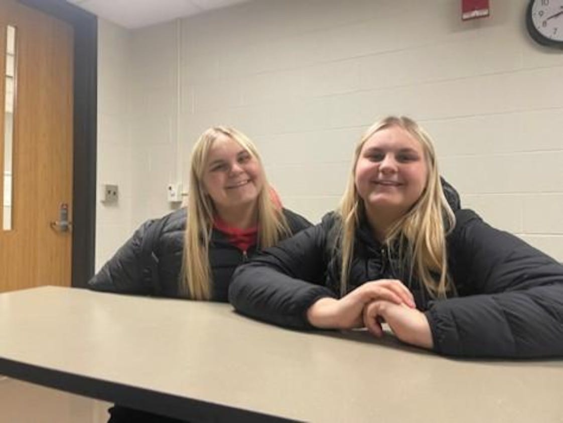 Twins Ella and Hannah Schultz sit in the back of a classroom, smiling, in matching black winter coats.