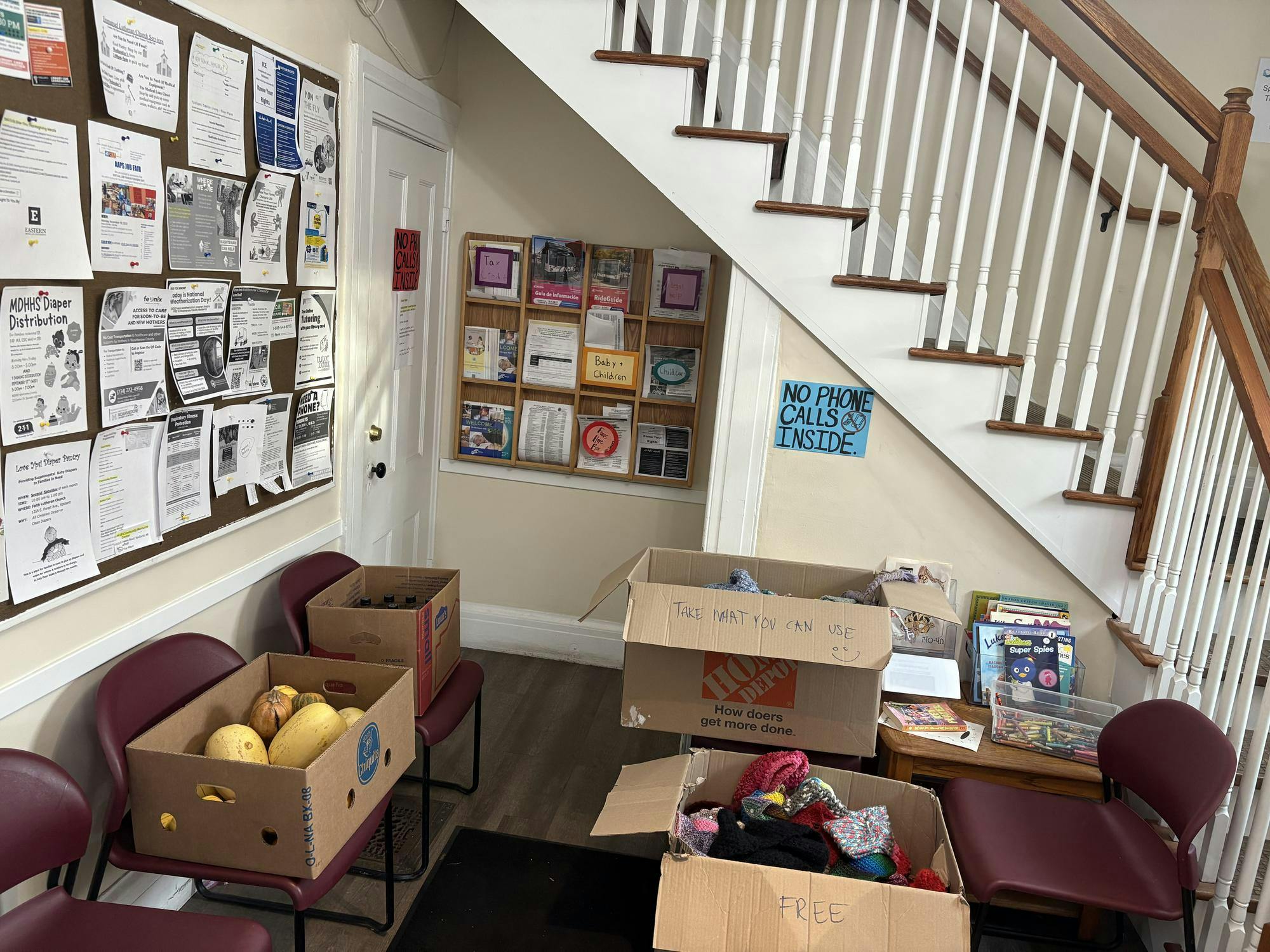 An image of a building lobby with flyers on the walls and bulletin boards, and chairs stacked with with boxes of squashes, food items, and clothing items. The boxes are labeled with "Free" and "Take what you can use."