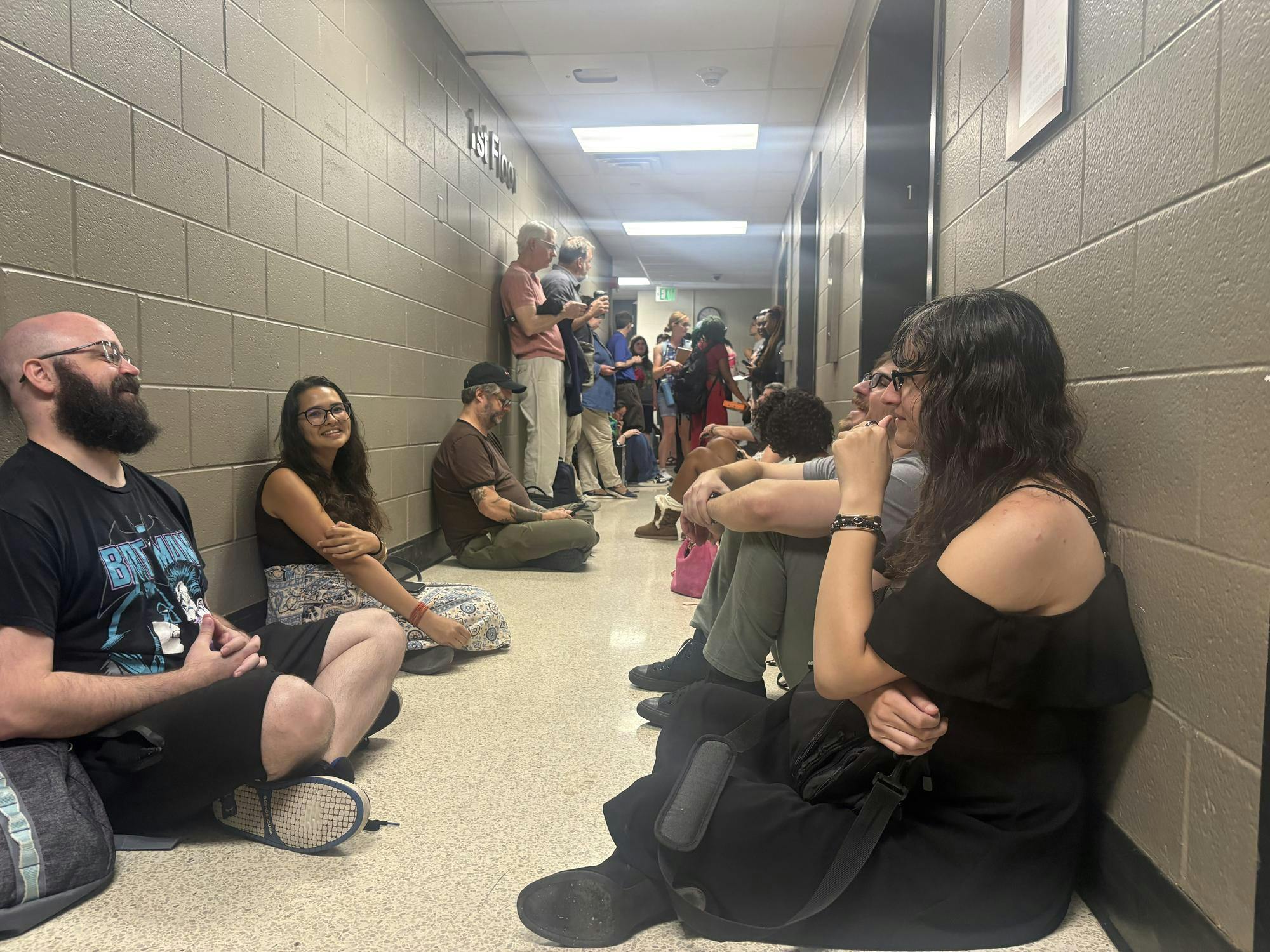 Several students sit on the floor against the concrete walls of the basement in Pray-Harrold, next to the elevators. More students and professors are seen in the background.