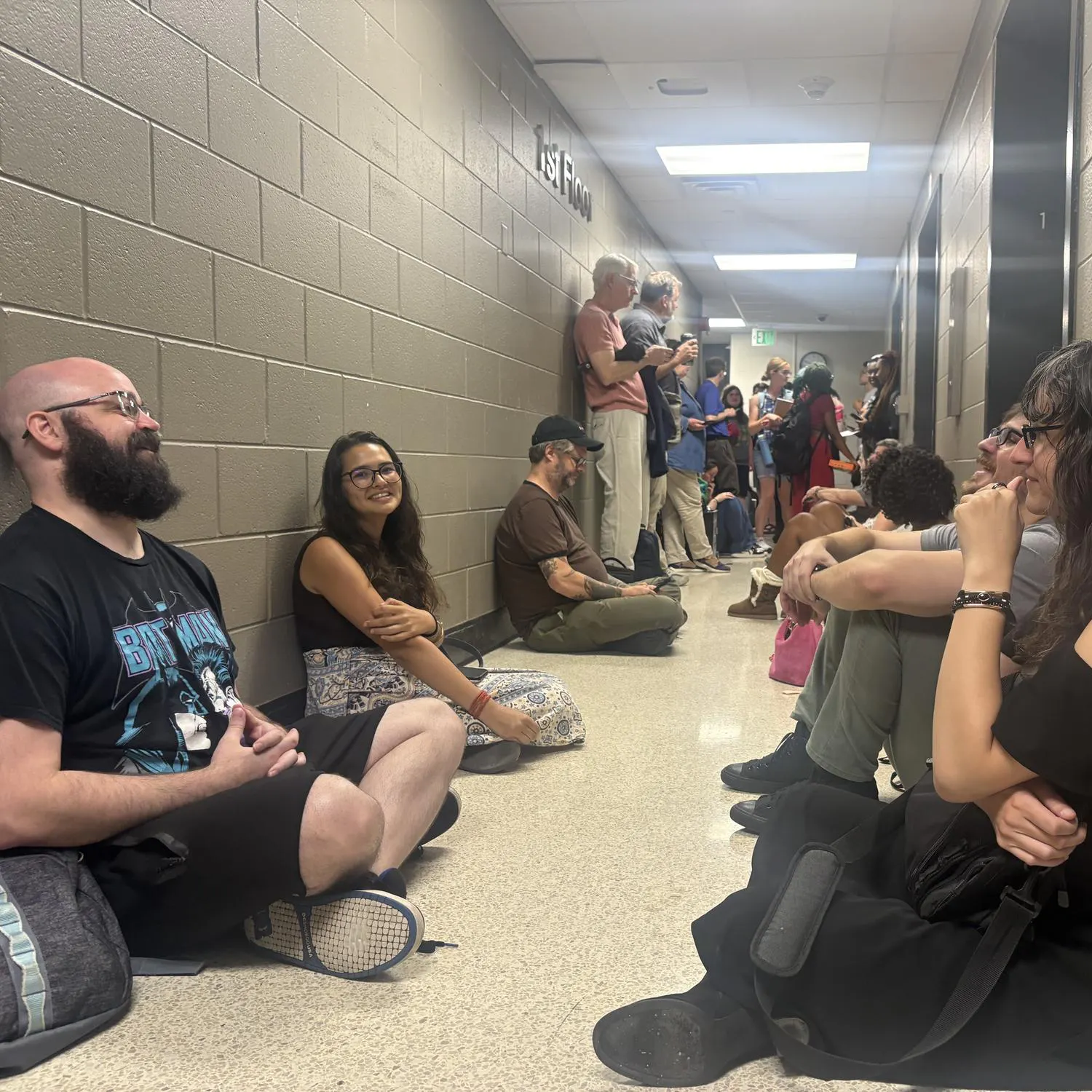 Several students sit on the floor against the concrete walls of the basement in Pray-Harrold, next to the elevators. More students and professors are seen in the background.