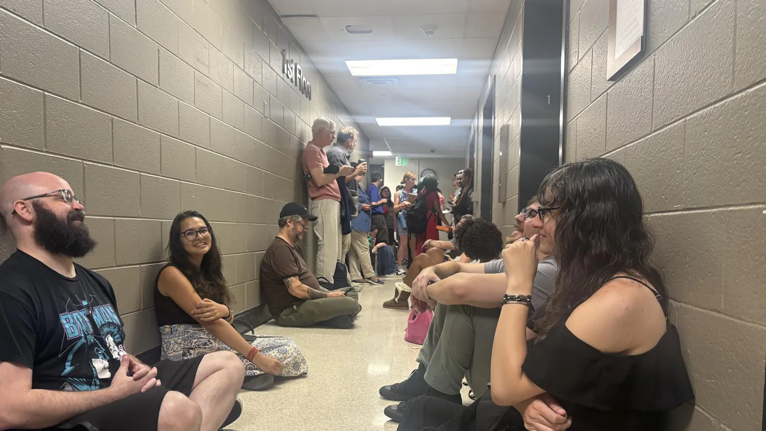 Several students sit on the floor against the concrete walls of the basement in Pray-Harrold, next to the elevators. More students and professors are seen in the background.