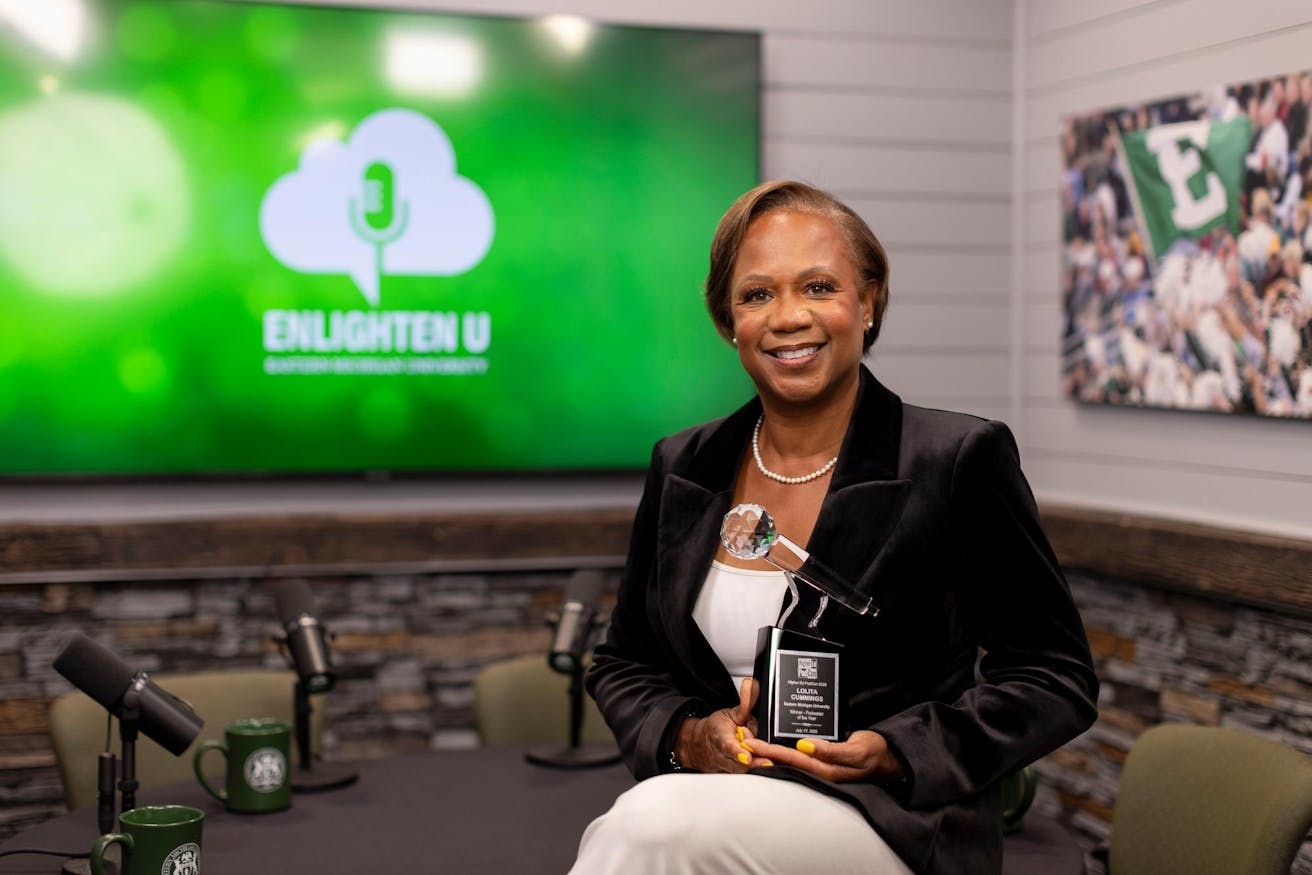 A woman holding an award that has her name on it, standing in front of a table with microphones on it and a green television screen in the background.