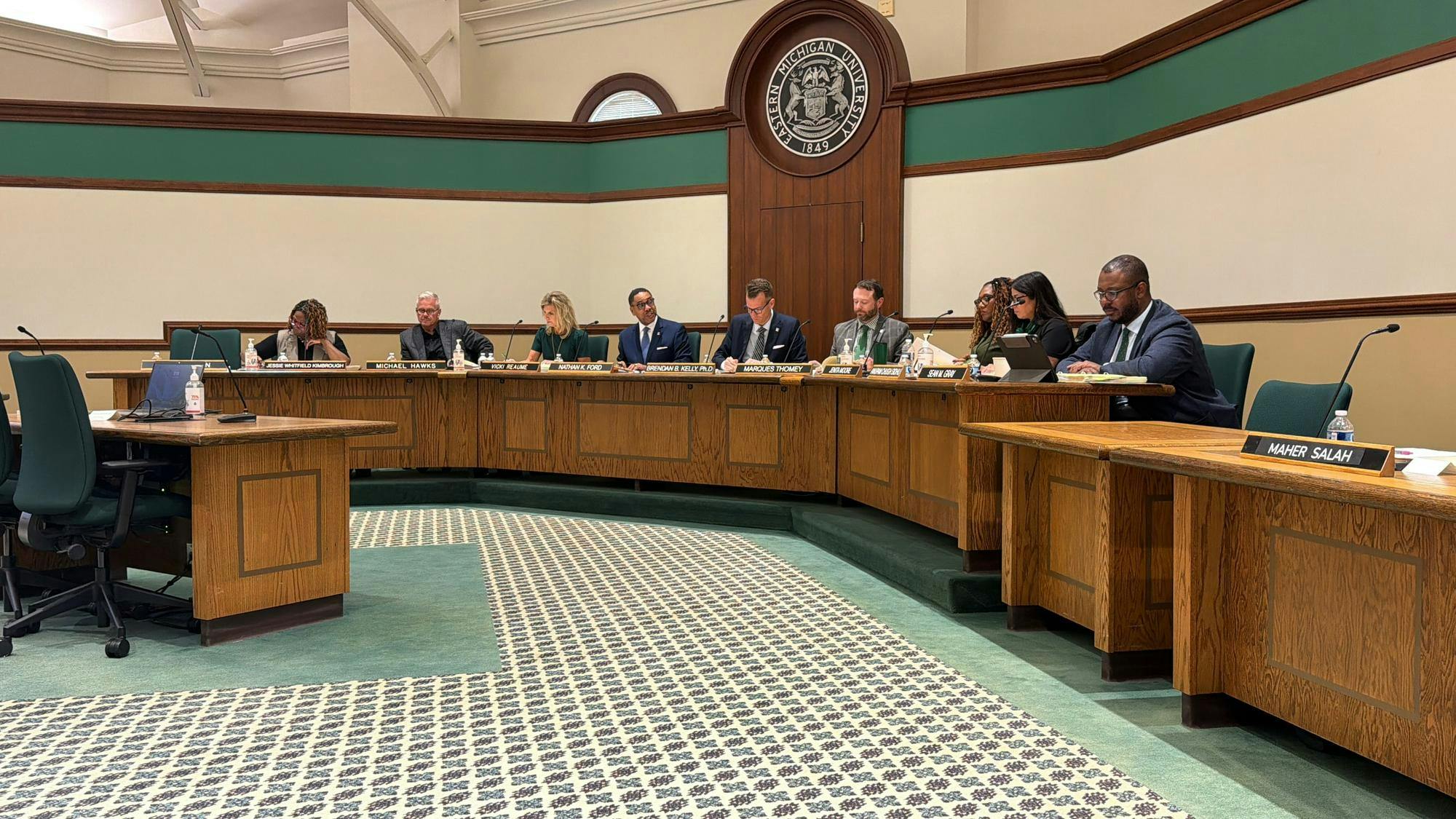 Brendan Kelly and the Eastern Michigan University Board of Regents sit behind a long desk with EMU's seal on the wall behind them in a large hearing room.