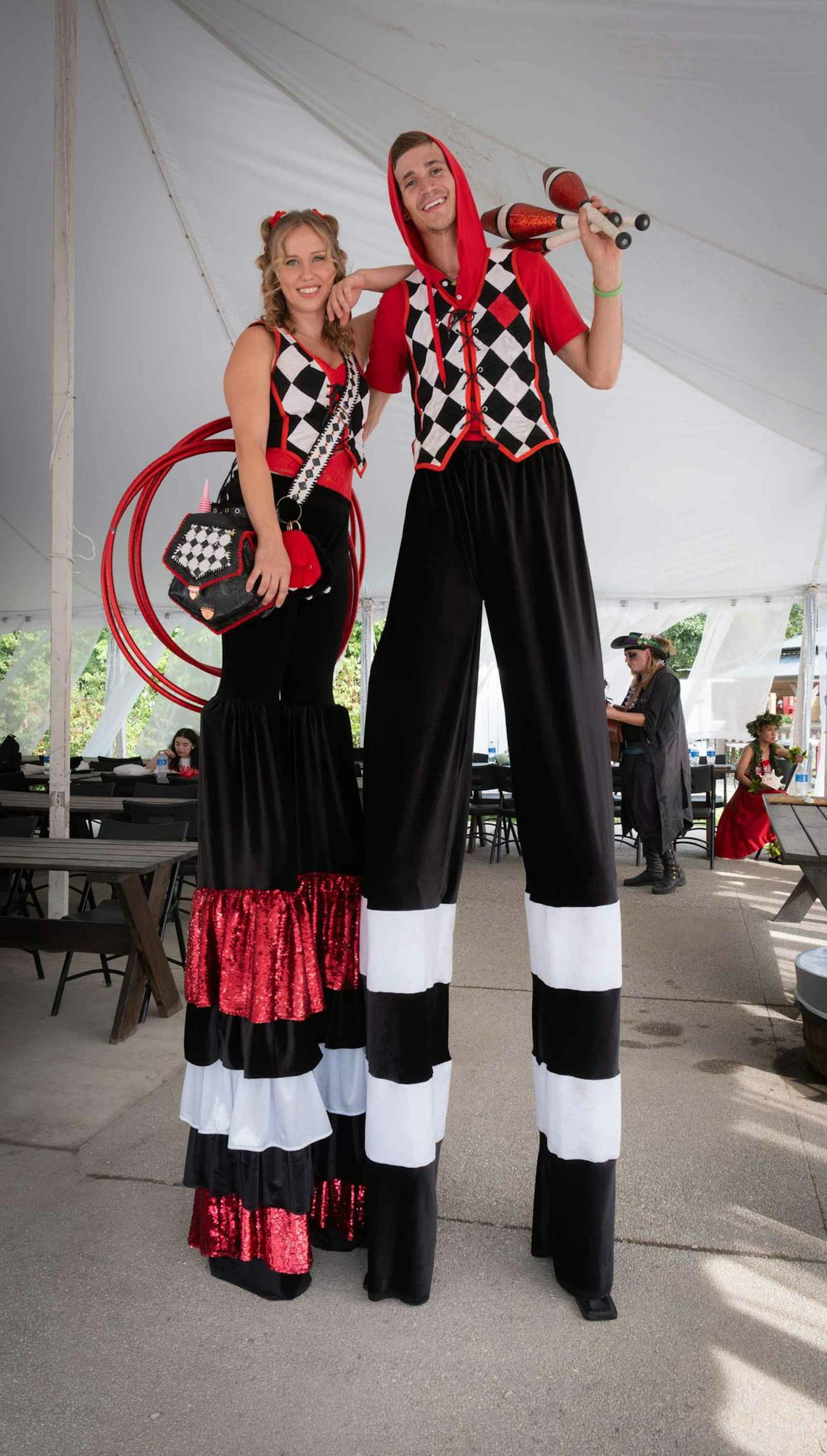 Two people stand on stilts with coordinating red, black and white outfits and holding props, such as hoops and juggling pins.