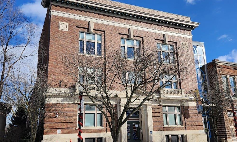 A large, rectangular-shaped brick building with several rows of windows. In front of the building is a tree that has lost all its leaves. The sky above is blue with a few white clouds, and there is another brick building to the right of the main building pictured. 